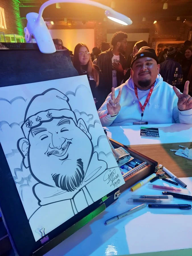 A caricature is on a table easel with markers strewn about and a smiling man in a white hoodie makes 2 peace signs in the background.
