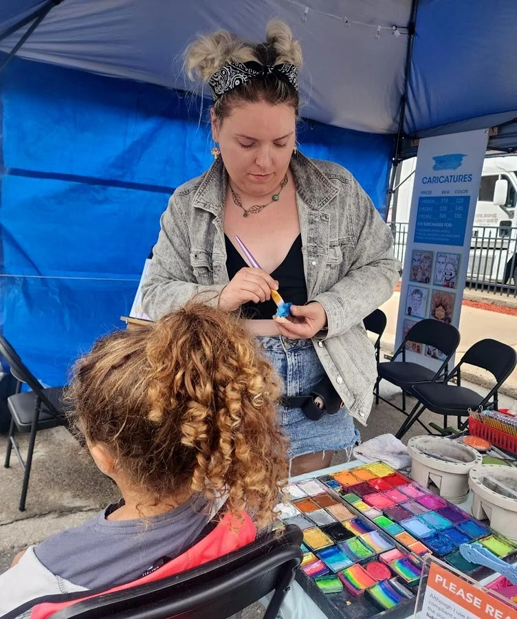 An artist is loading her brush and is about to apply paint to a child's face at a festival.