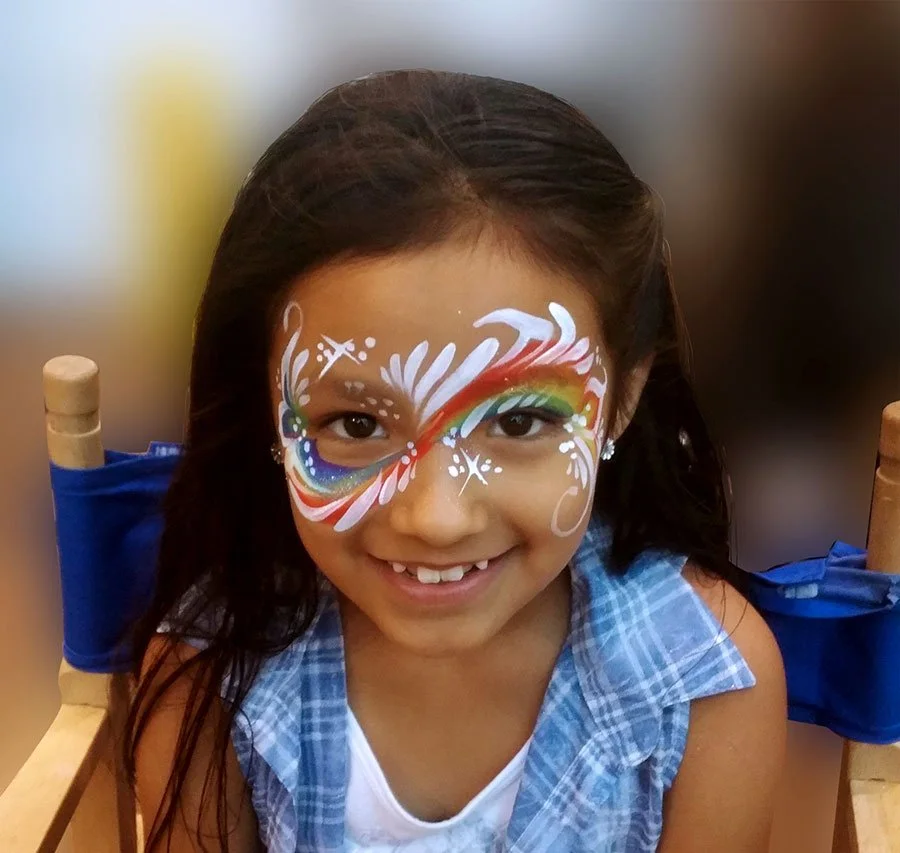 A smiling girl is wearing a rainbow face paint with white details.