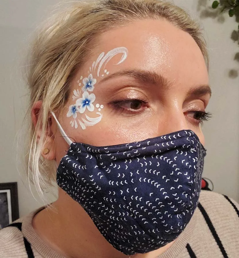 A masked woman wears a face paint of white and blue flowers and white swirls.
