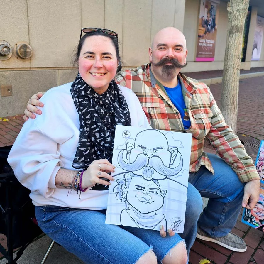A smiling woman with a scarf and pink cheeks and a bald man with a large handlebar mustache sits on the side of the road and holds their caricature.
