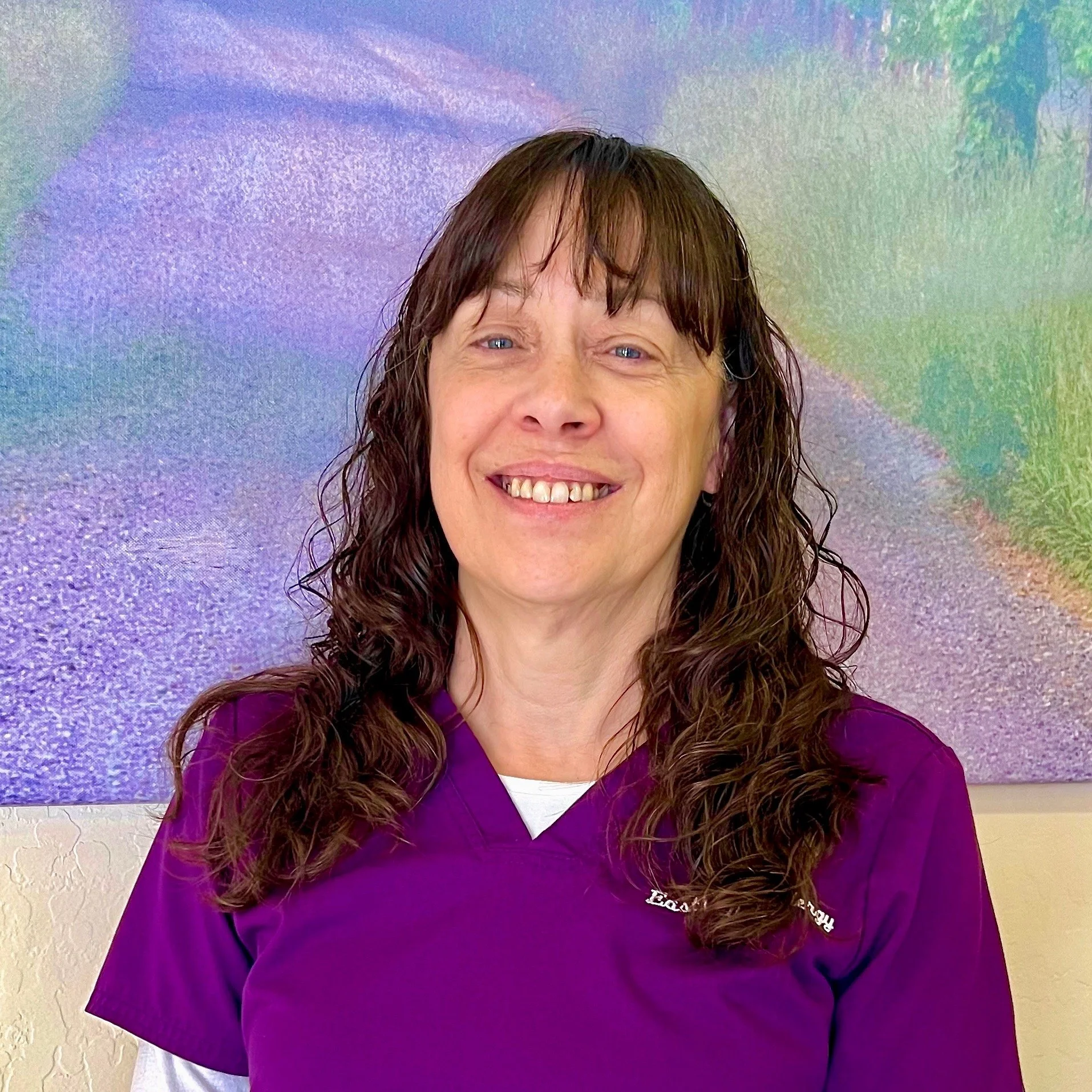 Smiling woman with brown curly hair wearing purple scrubs standing in front of a colorful abstract background.