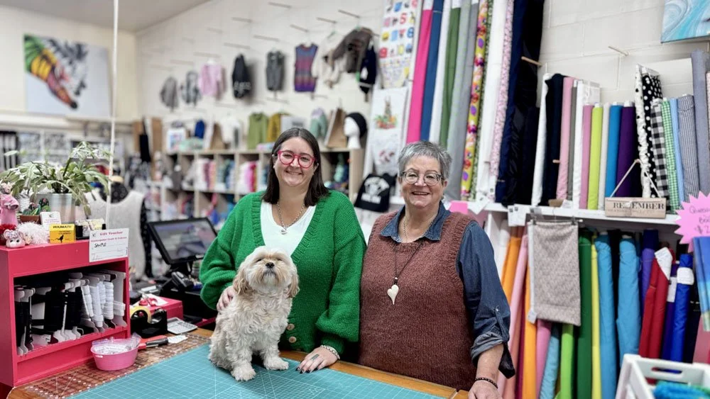 Two women smiling behind a checkout counter with a small dog, in a fabric and craft store with shelves of colorful fabrics and crafting supplies.