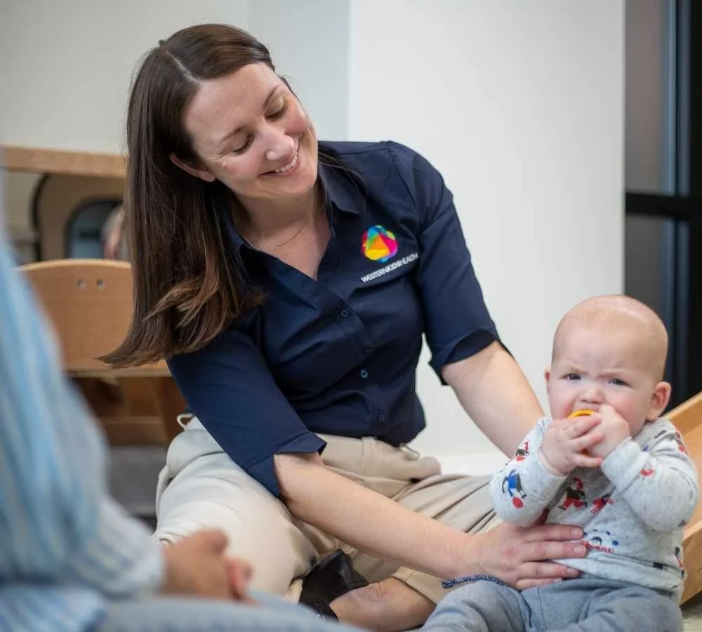 A woman with long brown hair wearing a navy blue shirt with a colorful logo is smiling while sitting on the floor with a young boy who is wearing a gray sweater with red and blue designs. The woman has her hand on the boy's stomach, and the boy is holding and chewing a yellow teething toy while looking at the camera.