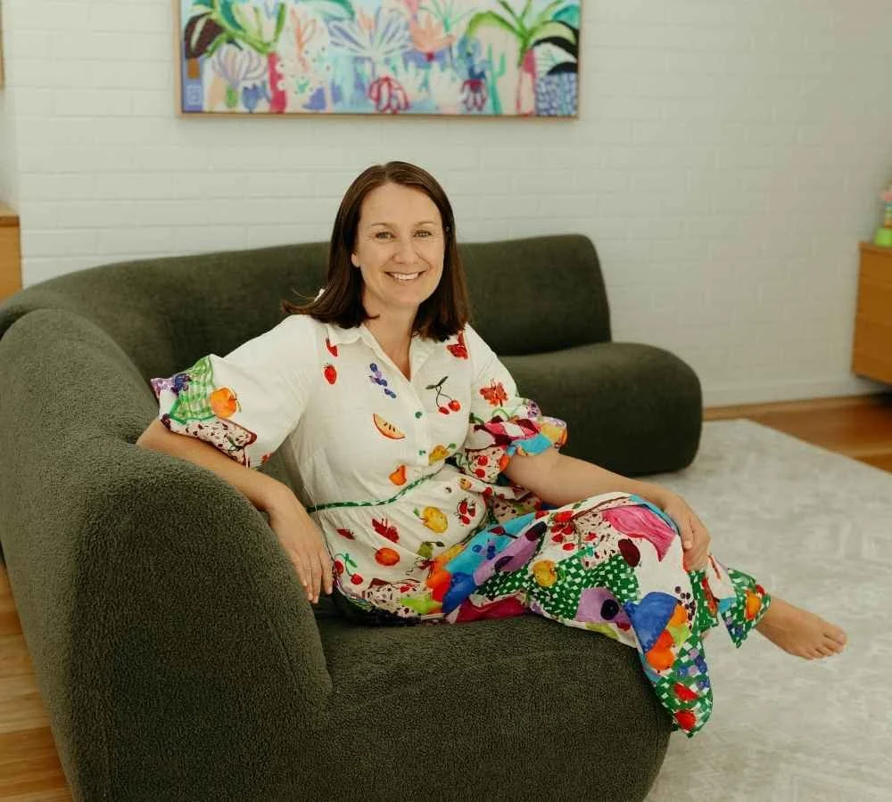 A woman with shoulder-length brown hair, smiling, sitting on a dark green sofa in a living room. She is wearing a white dress with colorful fruit and vegetable patterns. Behind her is a white brick wall with a colorful abstract painting hanging above the sofa. The room has light wood flooring and a light-colored area rug.