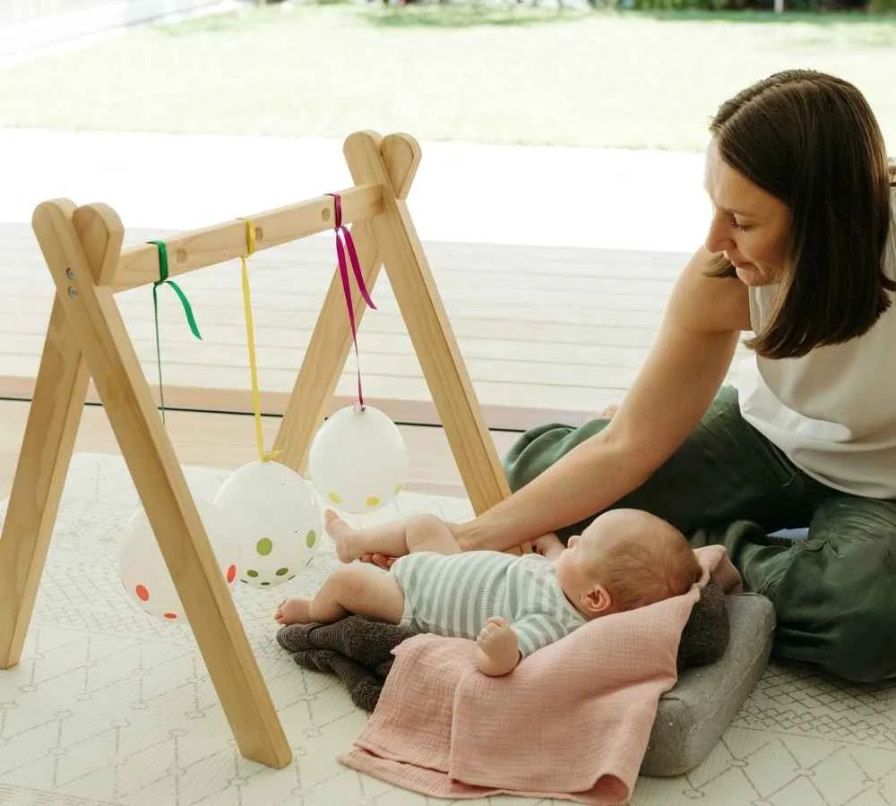 A woman playing with a baby on a soft blanket with hanging decorated egg-shaped balloons on a wooden stand.