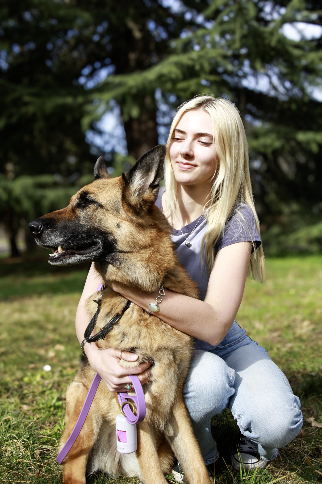 A young woman with blonde hair, wearing a gray shirt and light blue jeans, is kneeling outdoors on grass, hugging a large German Shepherd dog with a purple leash and a collar. The background features green trees and sunlight.