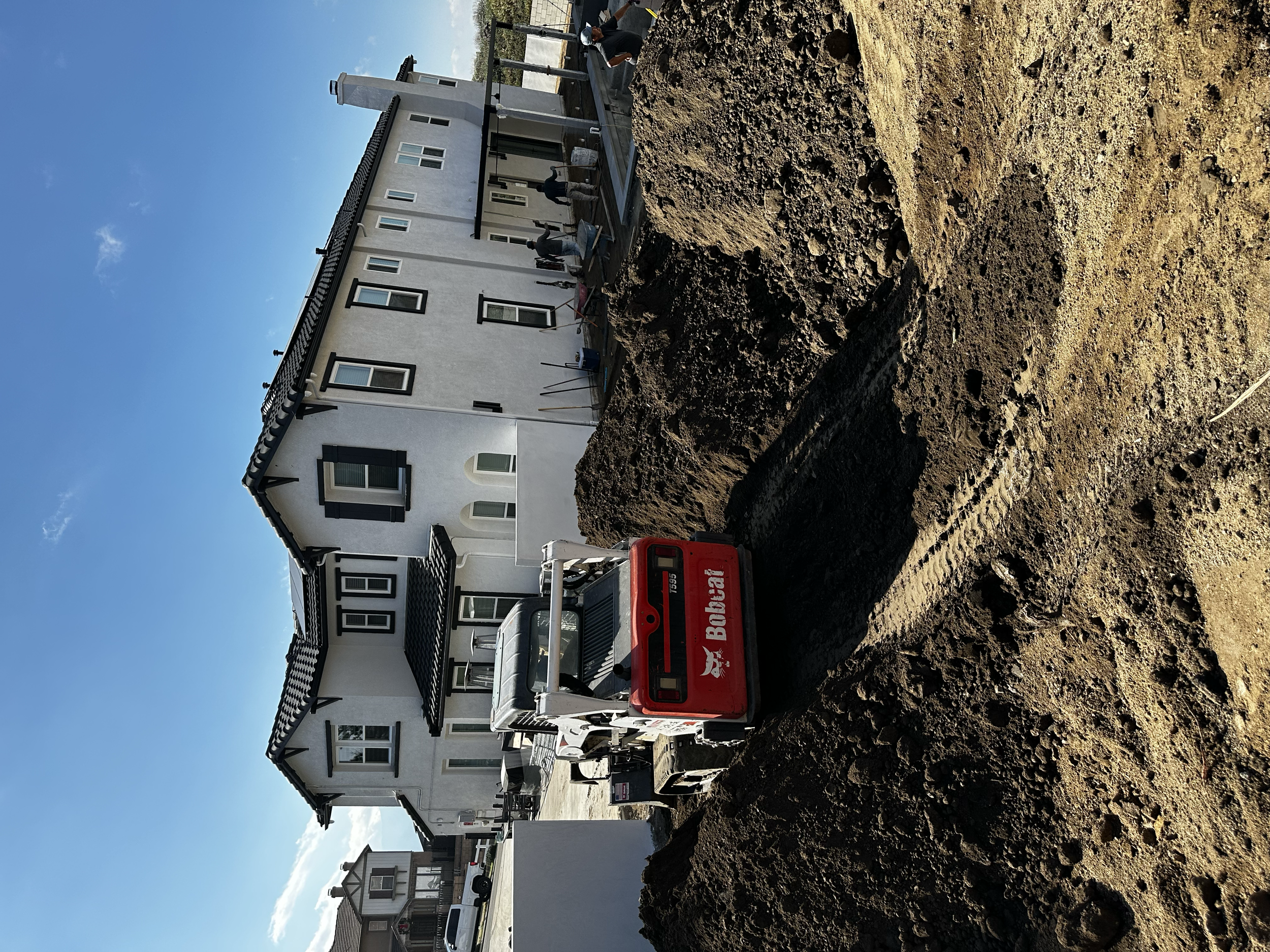 A construction site with a small excavator digging into the dirt in front of a white multi-story house with black window frames and a balcony, under a clear blue sky.