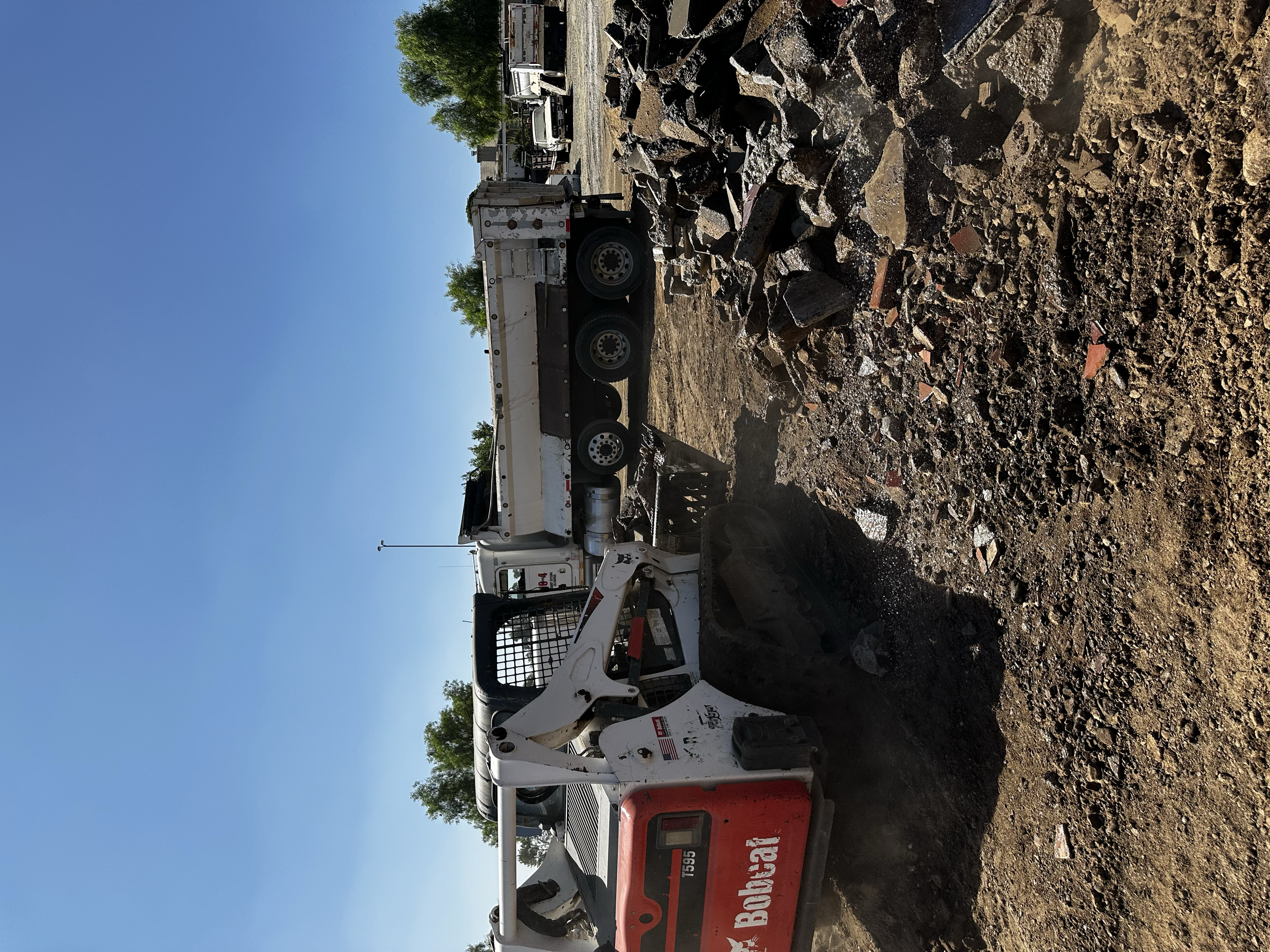 Construction site with a Bobcat skid-steer loader and a dump truck, dirt and rocks scattered on the ground, under clear blue sky with a few trees and parked cars in the background.