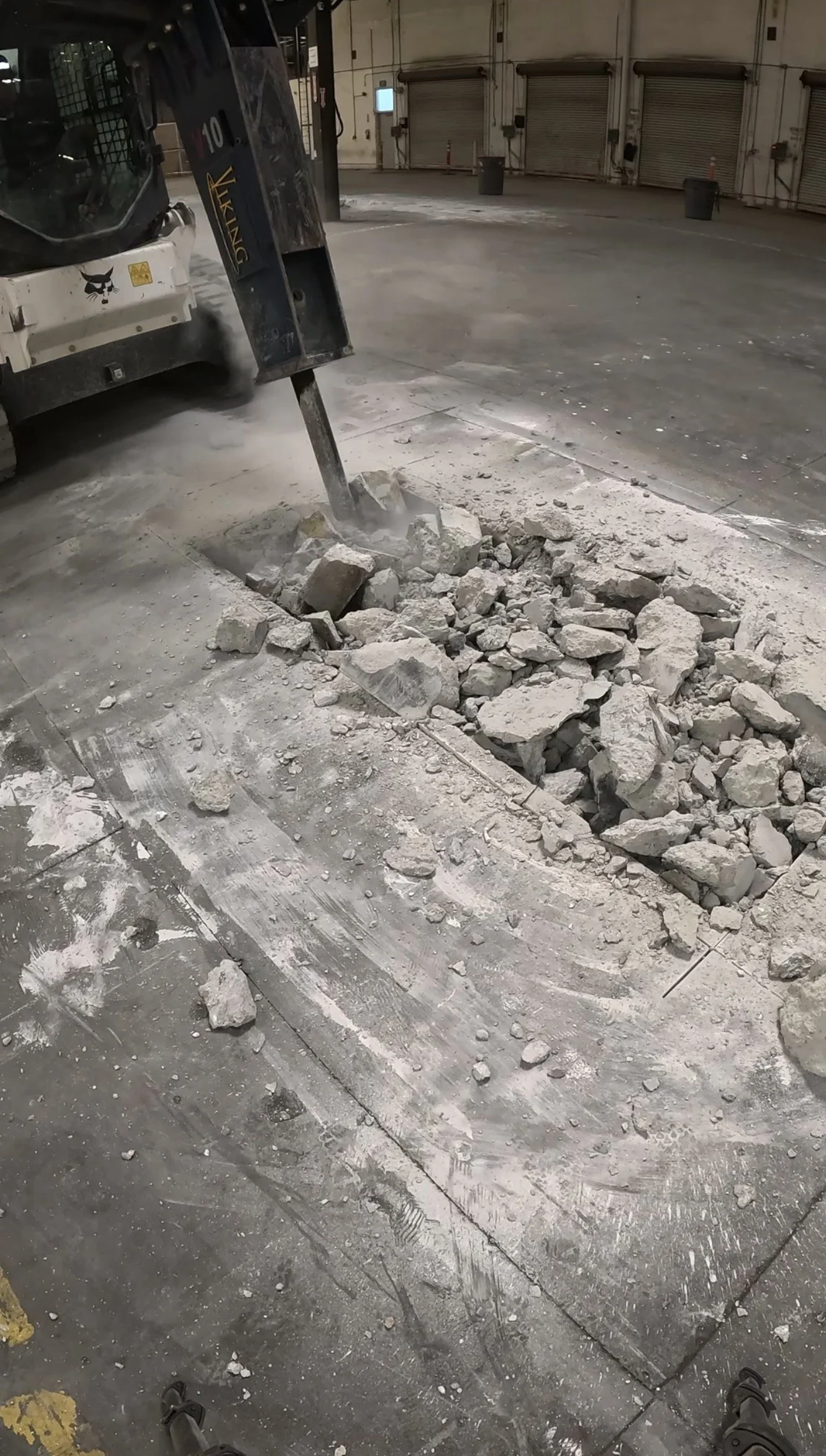 A construction worker using a jackhammer to break up concrete floor inside a large industrial garage or warehouse with closed garage doors in the background.