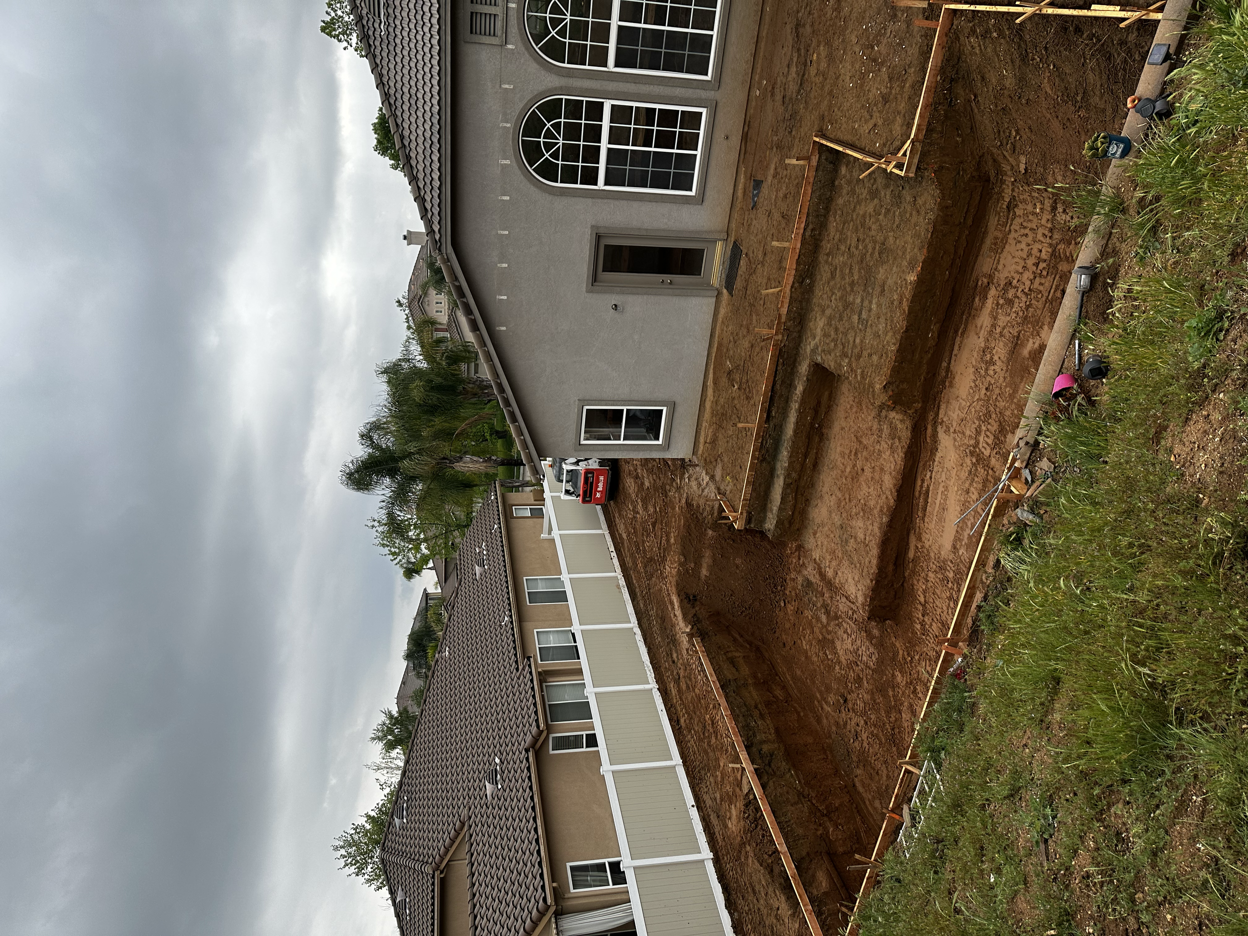 A residential backyard with an ongoing foundation excavation process. The image shows a cleared area of dirt, wooden framing for the foundation, and construction equipment in progress. Surrounding the site are neighboring houses and a cloudy sky.