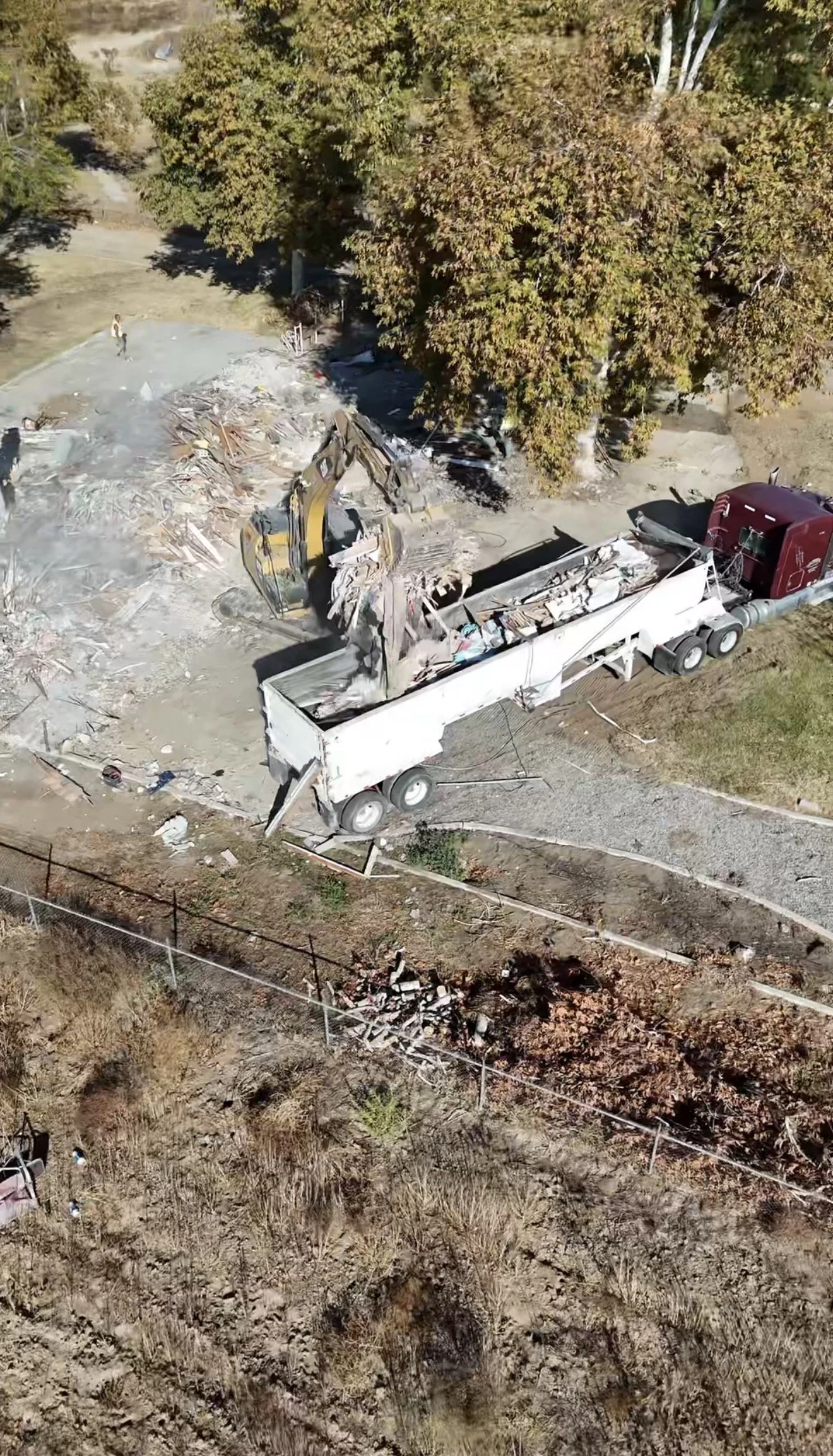 Aerial view of a construction site with a large truck removing debris with an excavator, surrounded by trees and dirt, and some workers present.