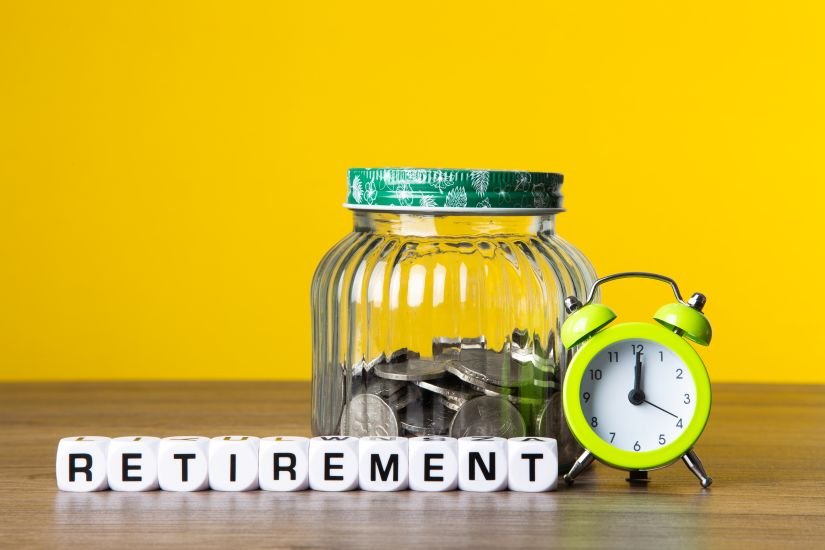 Jar of coins next to an alarm clock and wooden letter tiles spelling "retirement," representing time and savings