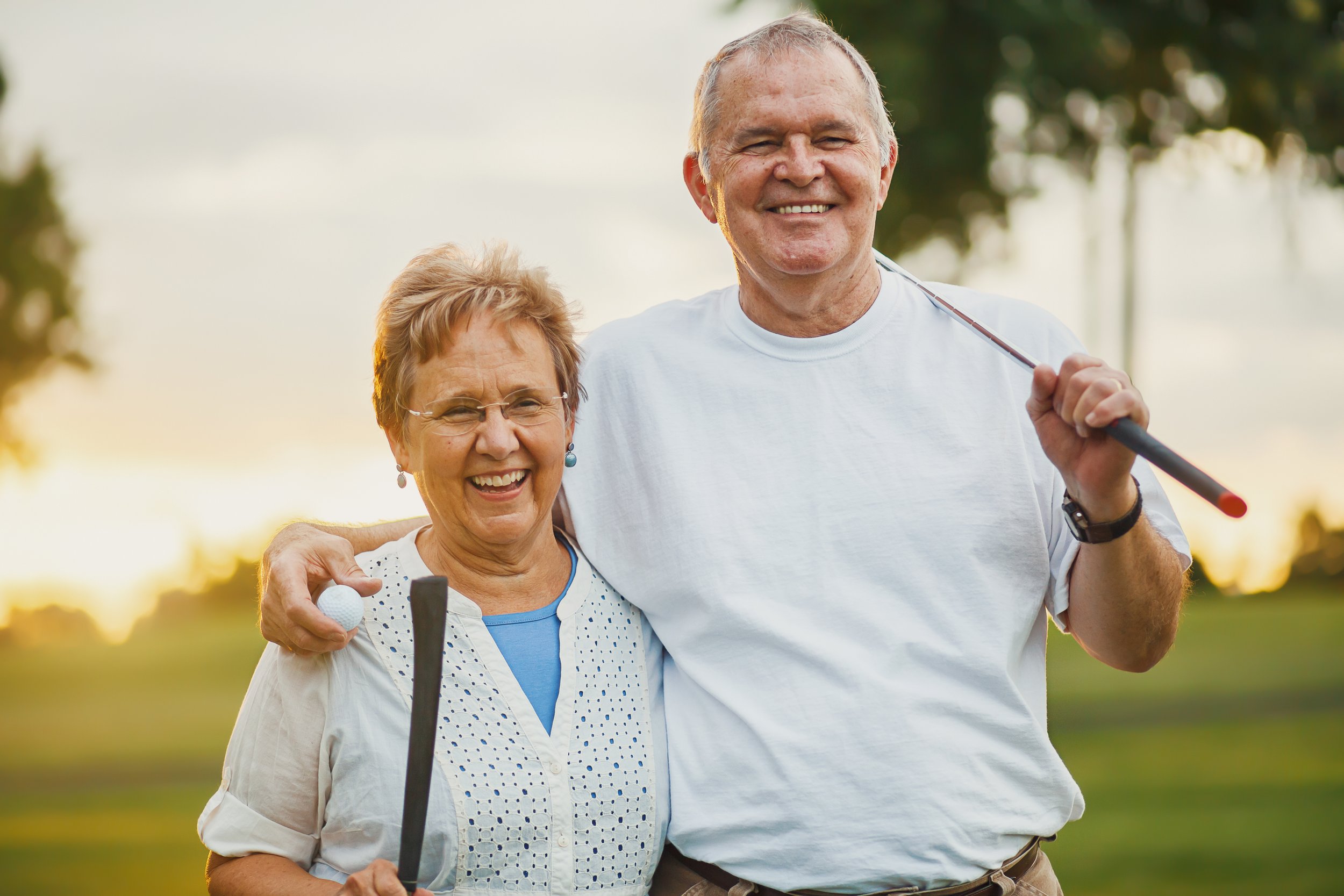 An elderly man and woman smiling and enjoying a day outdoors, holding golf clubs, with a blurred background of trees and sunlight.