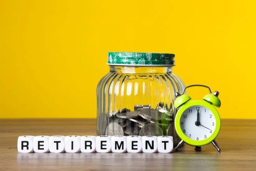 Jar of coins next to an alarm clock and wooden letter tiles spelling "retirement," representing time and savings