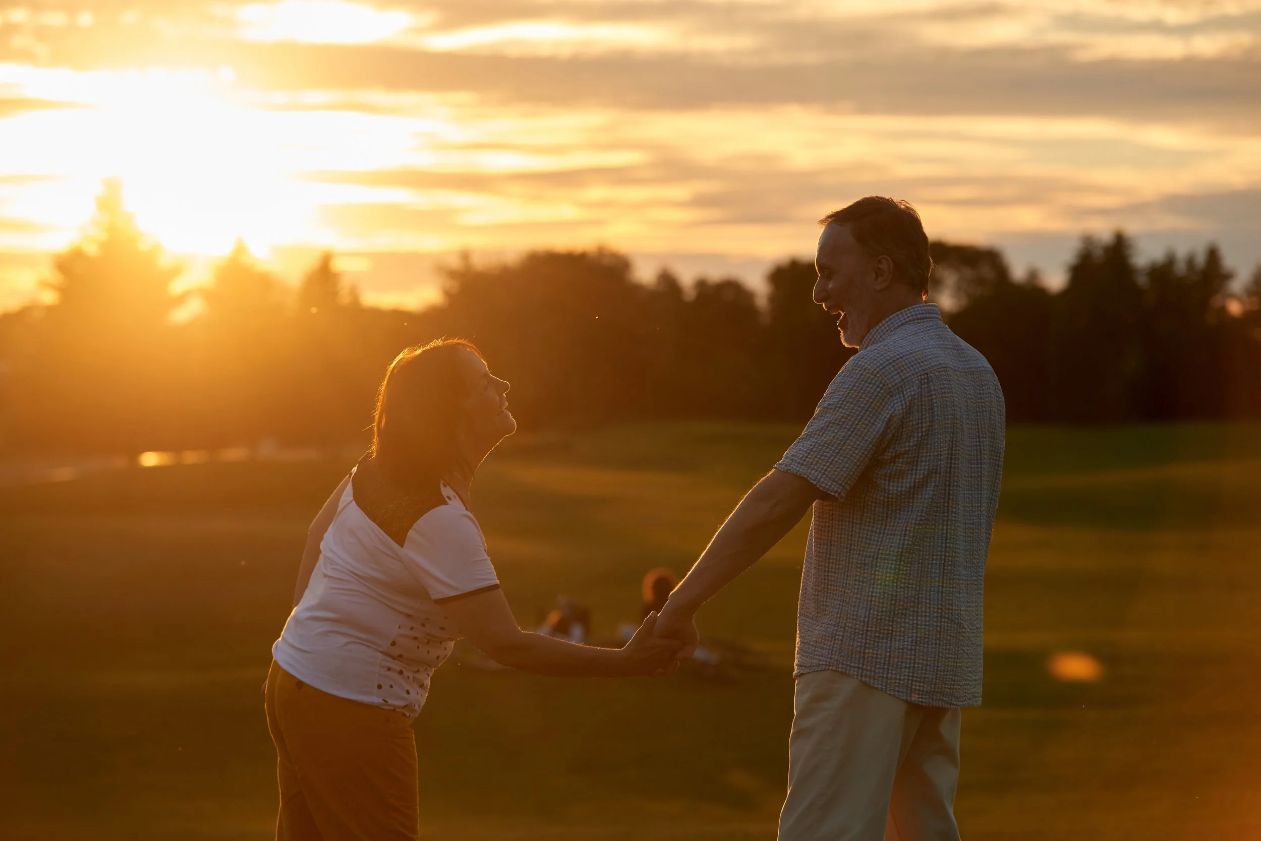 A man and a woman holding hands and smiling at each other during sunset in an outdoor setting, with trees and a cloudy sky in the background.