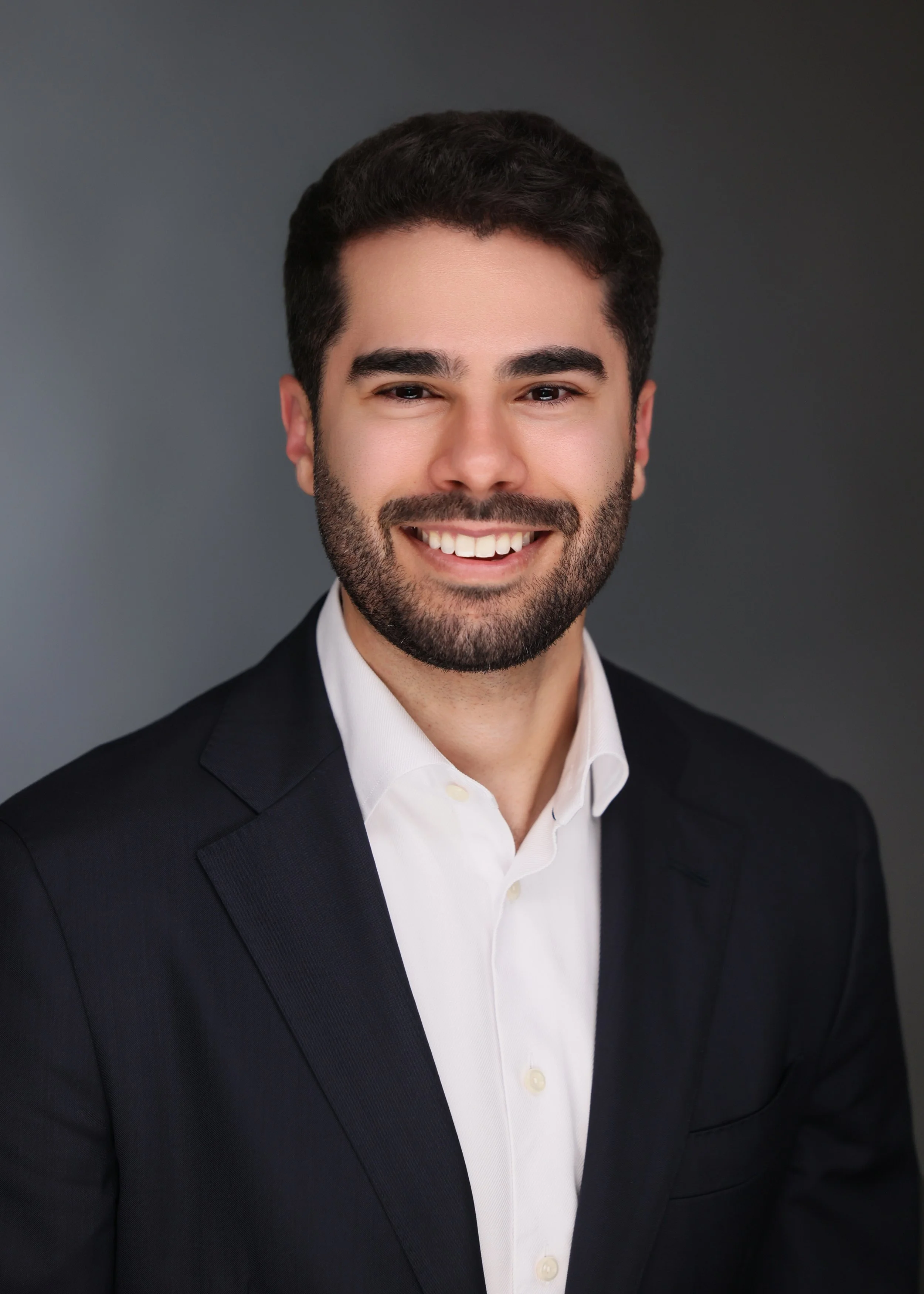 A man with dark hair and a beard, smiling, wearing a black suit and white shirt, against a gray background.