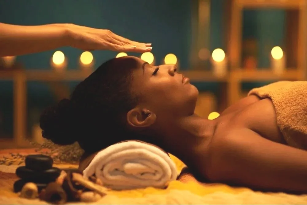 A woman lying face-up on a massage table, receiving a head massage in a spa or massage room, with lit candles and relaxing decor in the background.