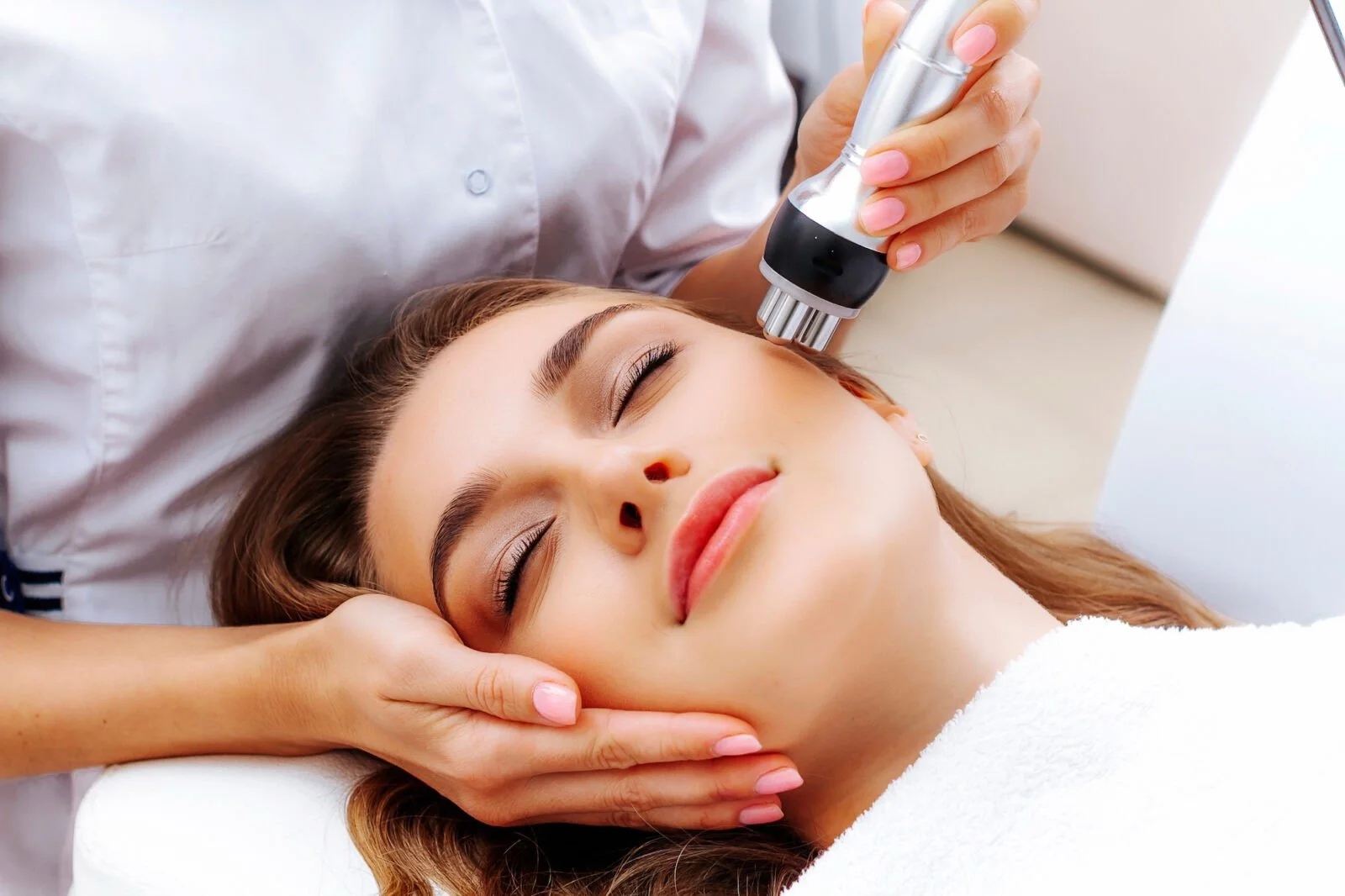 A woman lying down with eyes closed receiving a facial treatment using a small device on her face, with a professional in a white coat operating the device in a clinical setting.