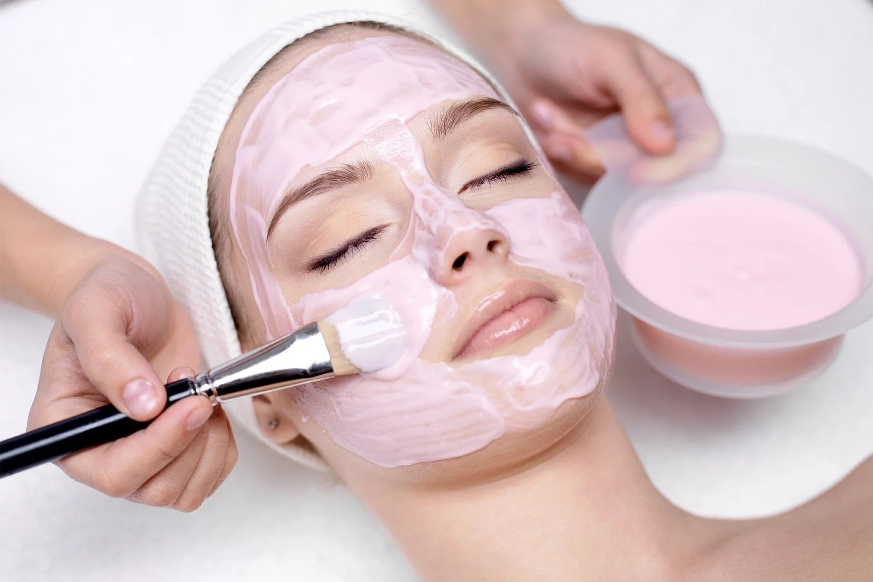 A woman receiving a facial treatment with a pink mask being applied with a brush by a spa technician.