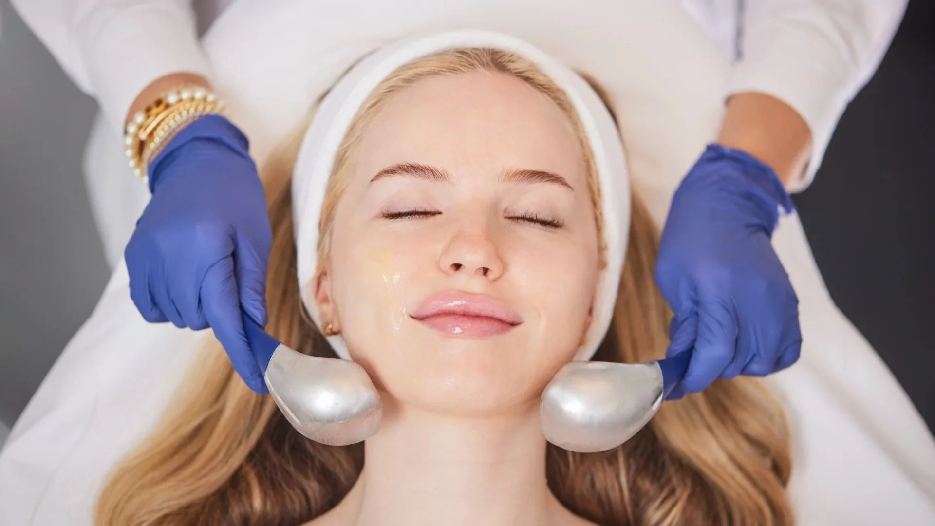 Woman receiving a facial treatment in a spa or dermatologist's office, lying down with eyes closed, with a therapist holding a metallic facial tool and wearing blue gloves.