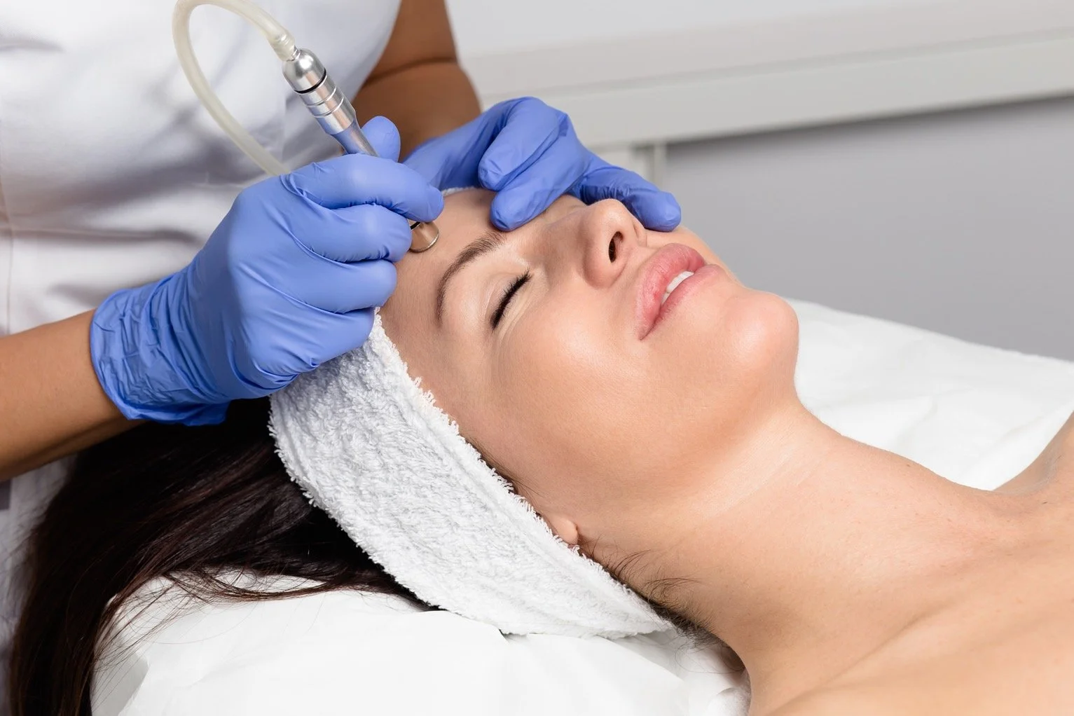 A woman receiving a facial treatment while lying down with her eyes closed, a towel around her head, and a healthcare professional wearing blue gloves performing a procedure with a handheld device.