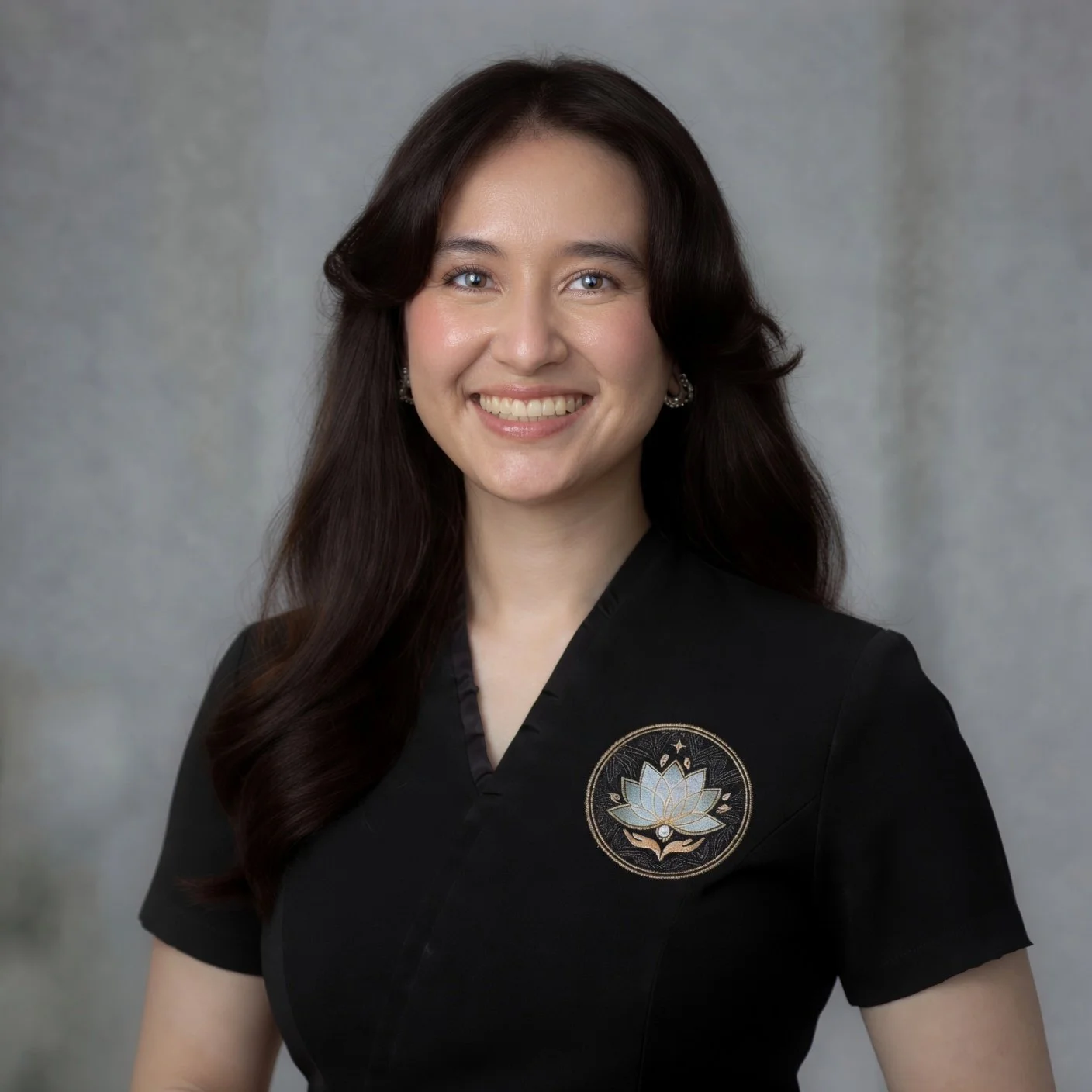 A woman with long dark hair, wearing a black top with a lotus flower emblem, smiling against a gray background.