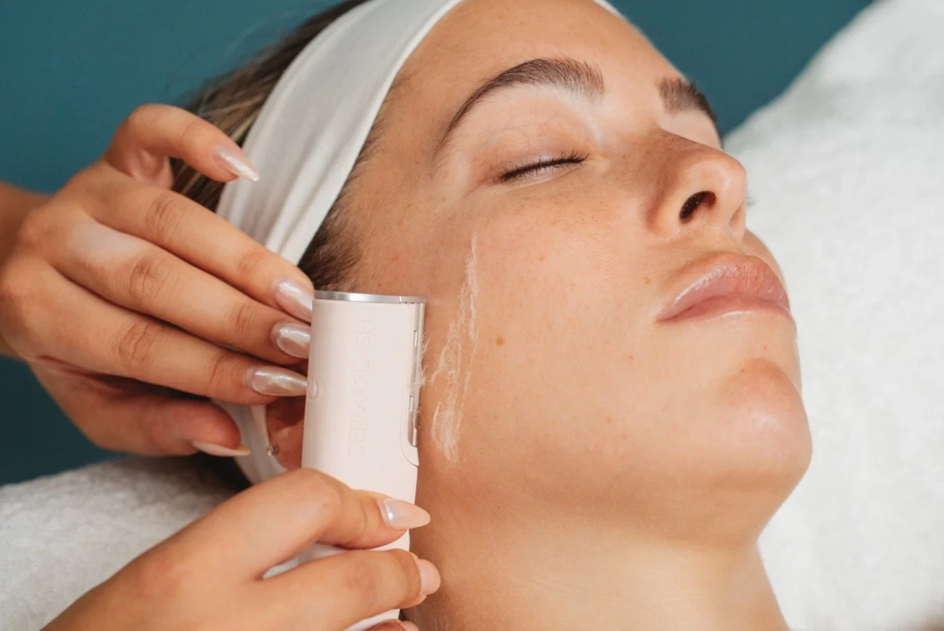 A woman receiving a facial treatment with a device applied to her cheek while lying down with her eyes closed.