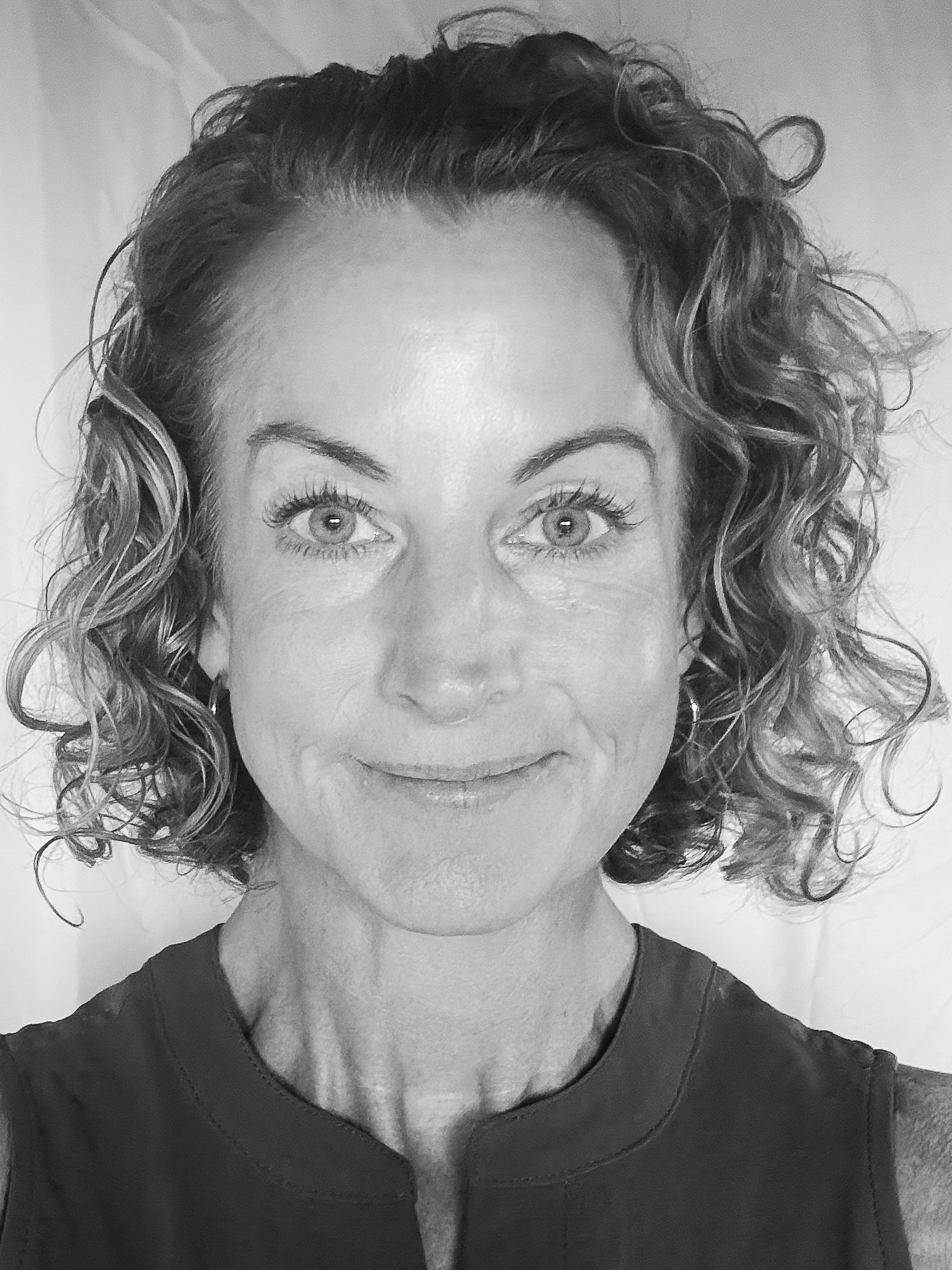 A close-up black and white portrait of a woman with curly hair, smiling slightly, wearing a sleeveless top.