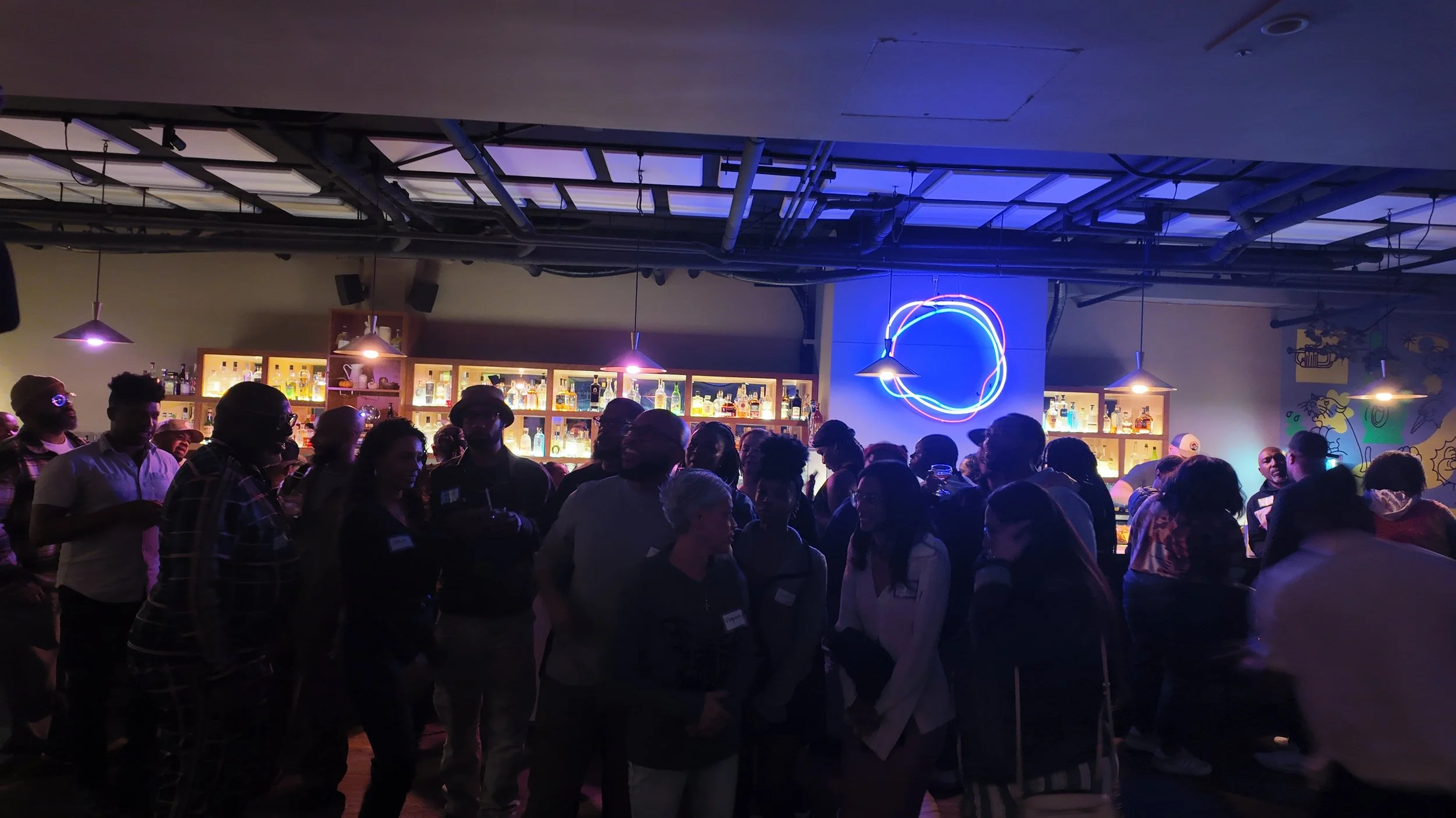 A crowded bar with people socializing, neon lights, and a bar with various bottles of alcohol in the background.