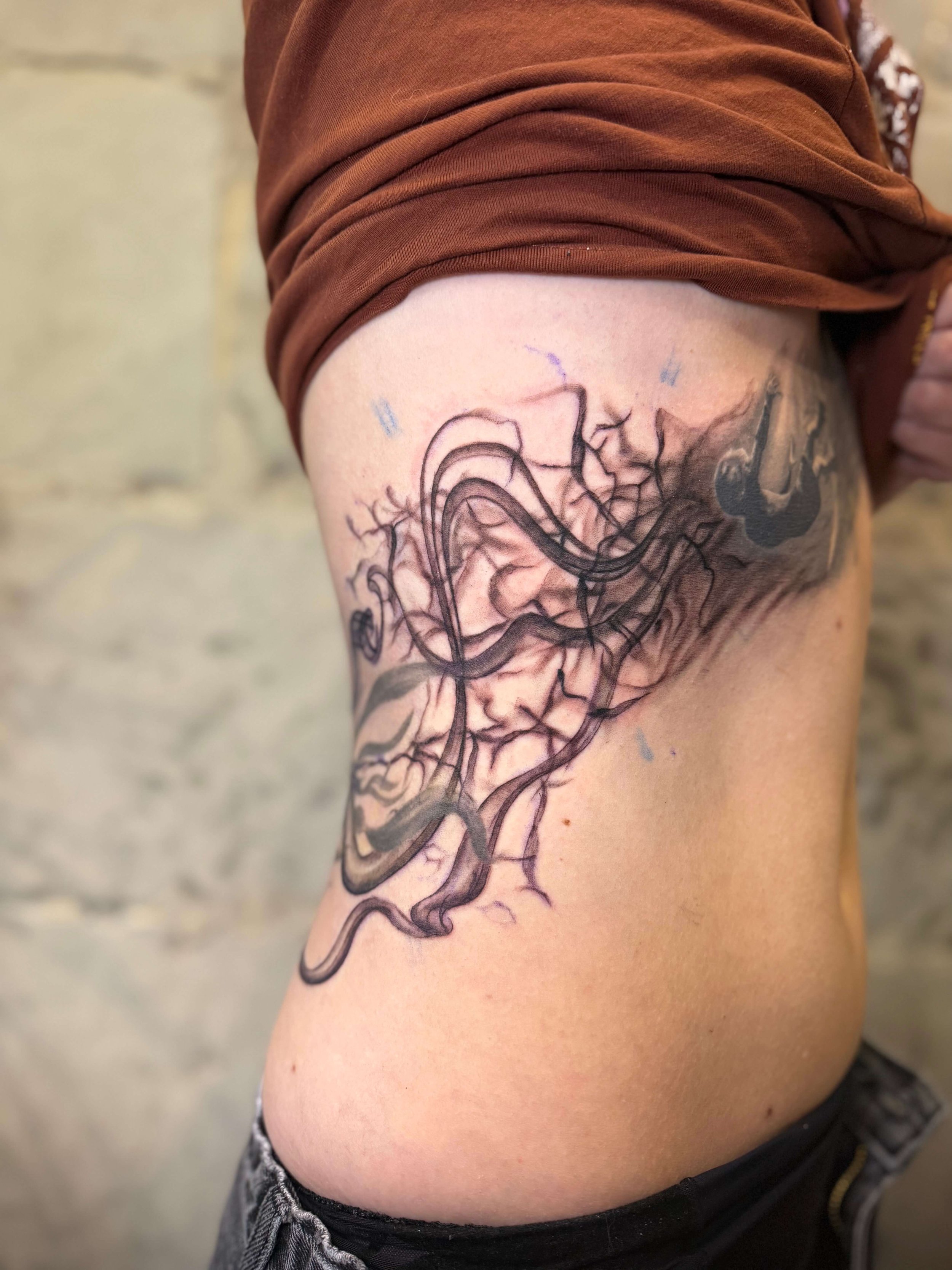 A tattoo of a detailed, abstract black ink design on a person's left side torso, featuring swirling and branching patterns.