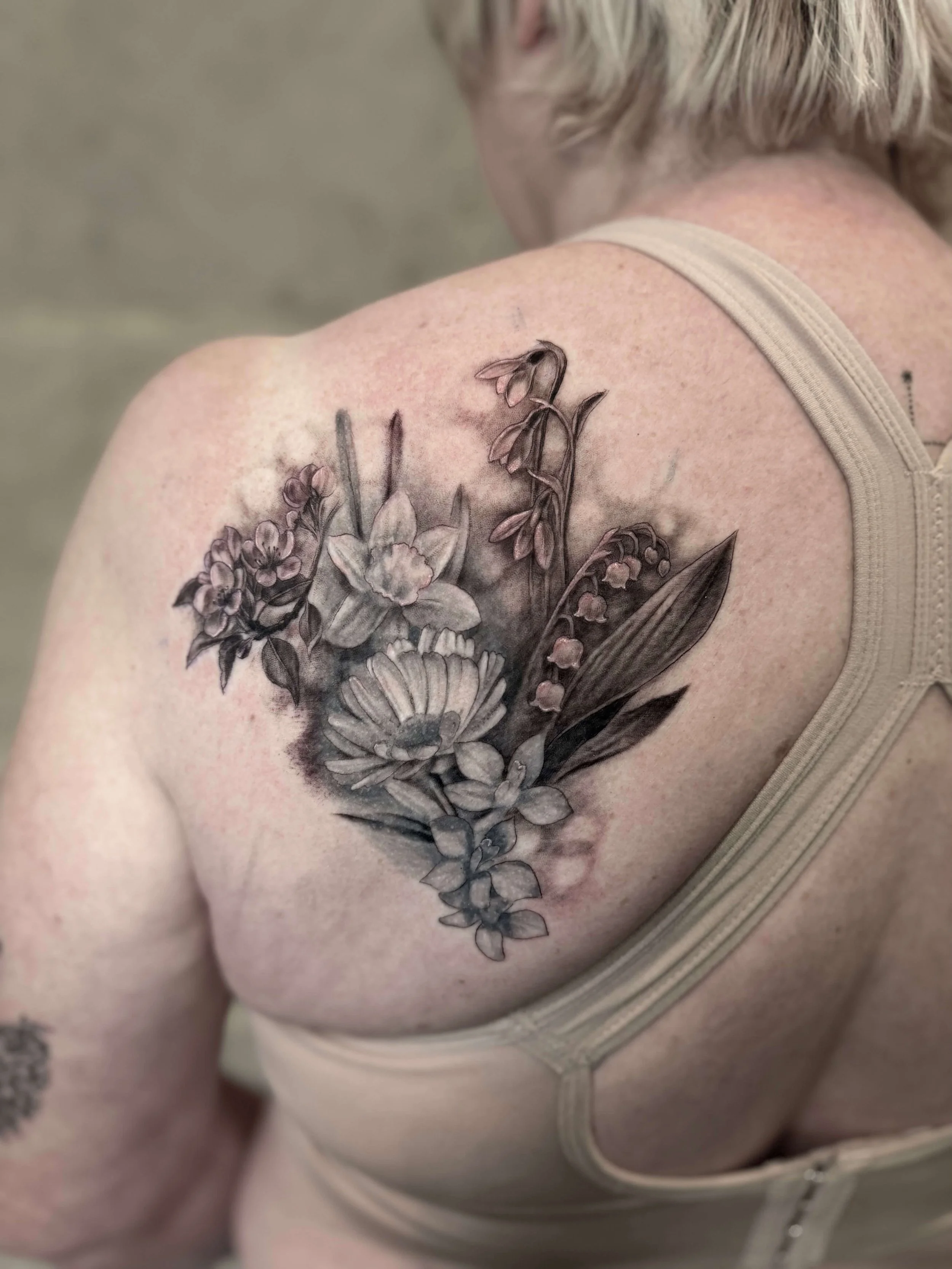 Black and gray floral tattoo on woman's shoulder featuring various flowers and leaves.