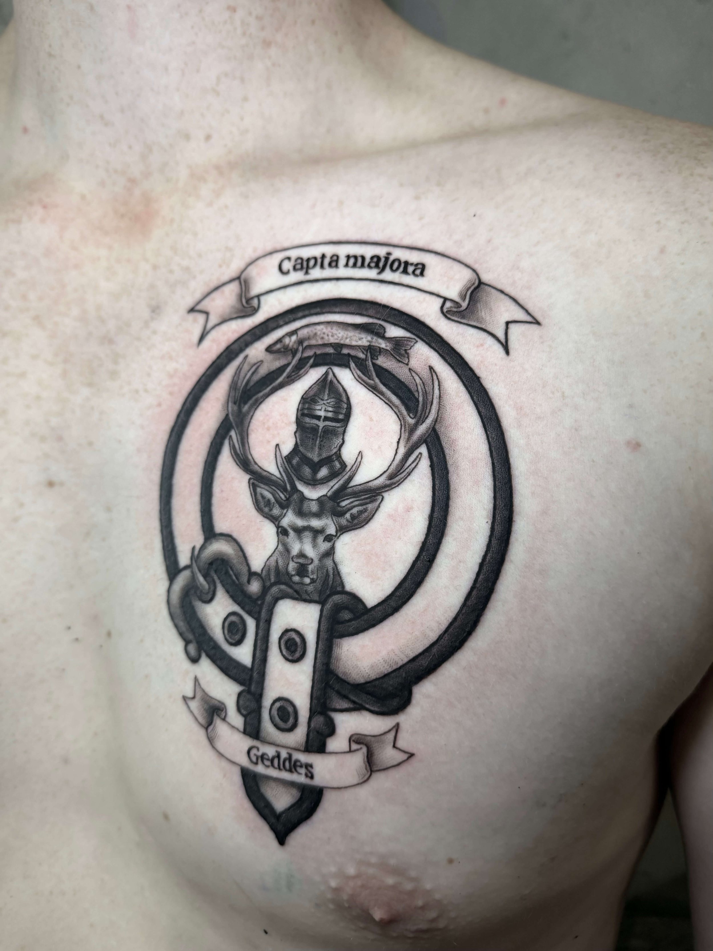 A tattoo on a person's chest featuring a stag's head with large antlers, a knight's helmet above it, and banner text reading "Captamajora" and "Geddes."
