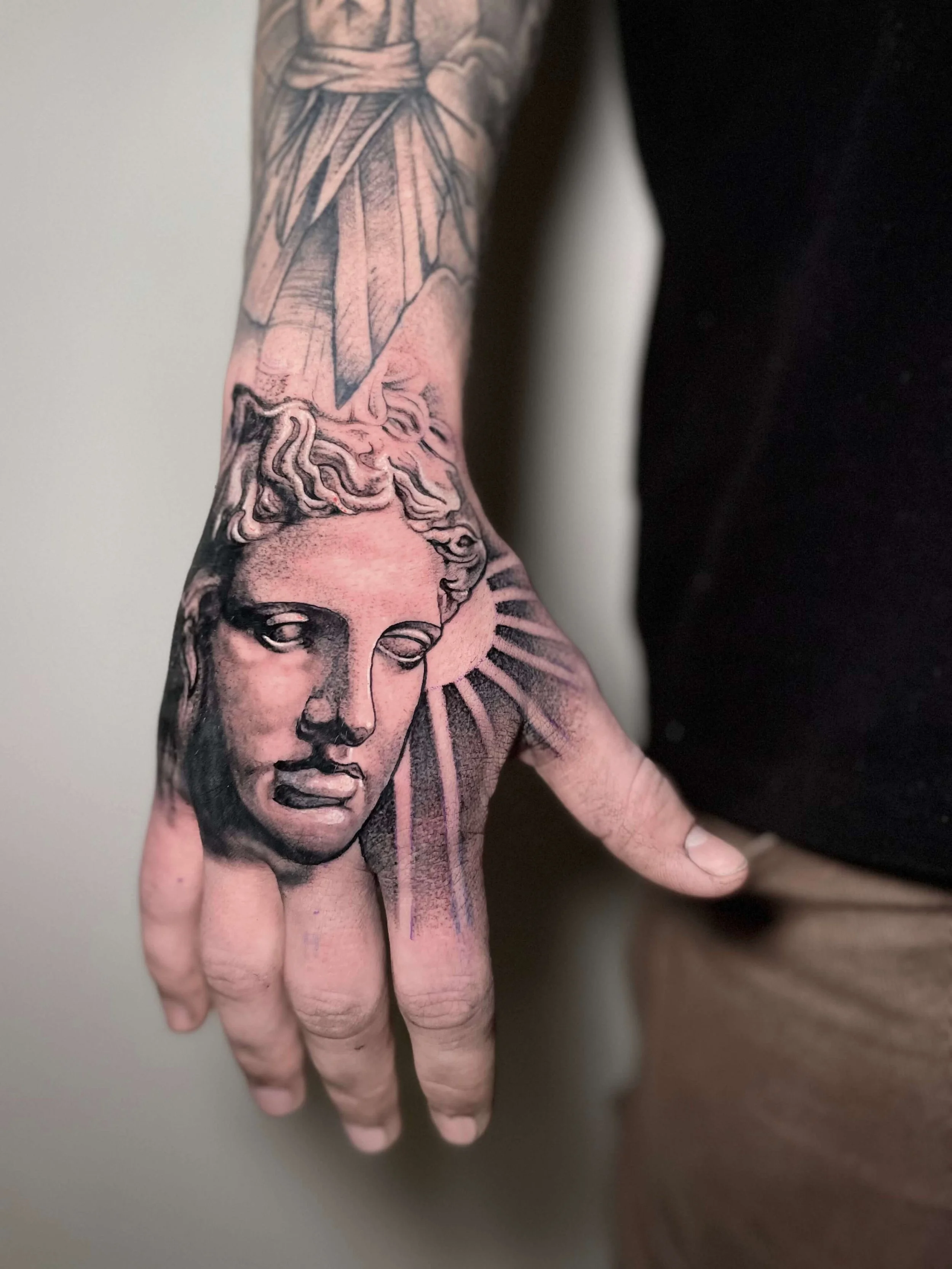 Tattoo of a classical sculpture of a woman's face with flowing hair on a person's hand and forearm, with rays emanating from behind the head.