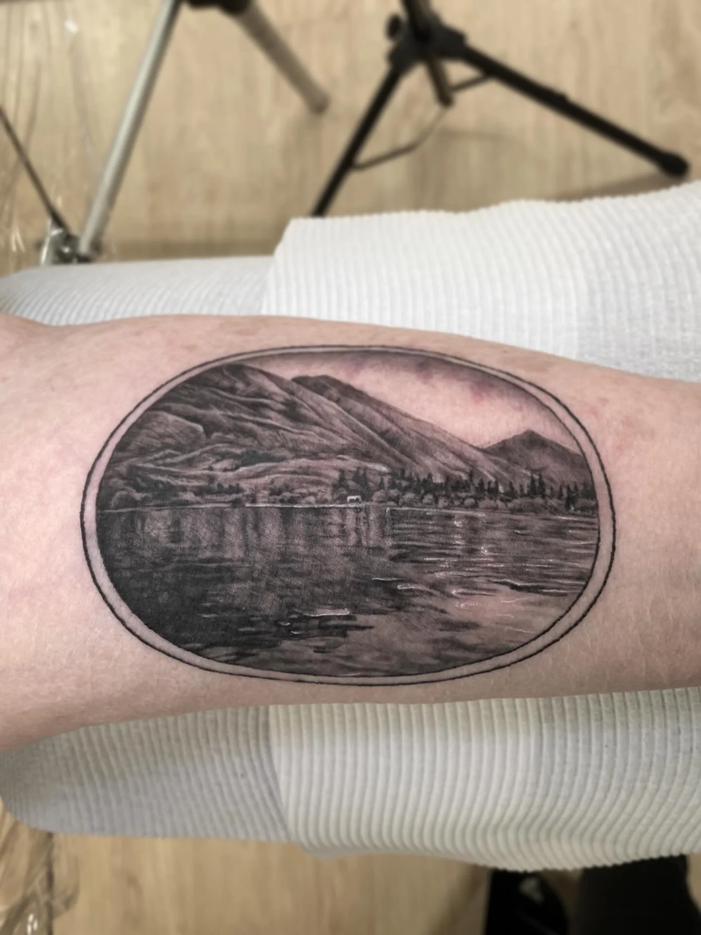 Black and gray tattoo of a mountain landscape with a lake and trees, enclosed in an oval frame on a person's arm.