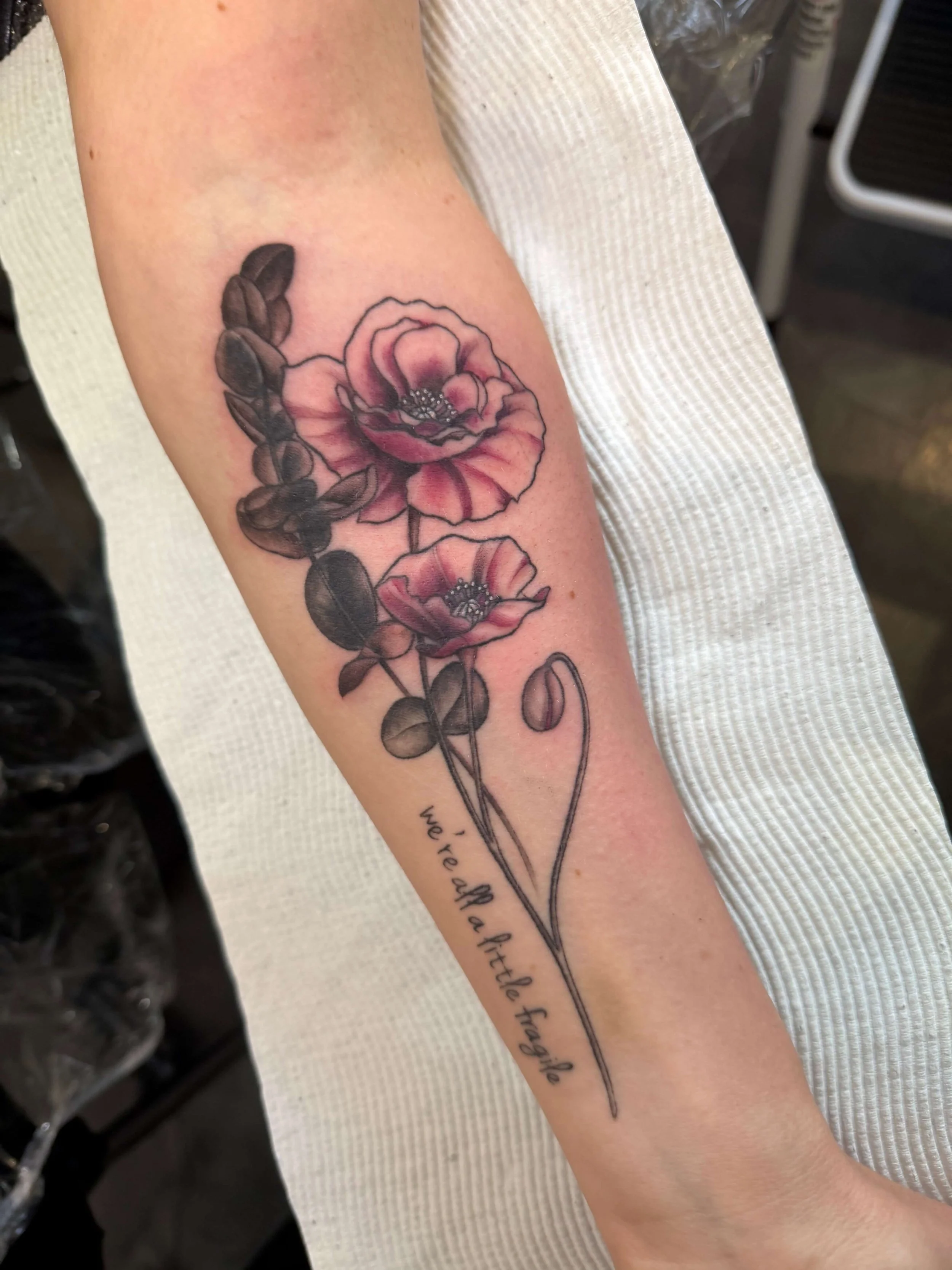 Tattoo of two pink flowers with green leaves and stems on a person's arm, with the words "we lift a little flag" written in cursive below the flowers.