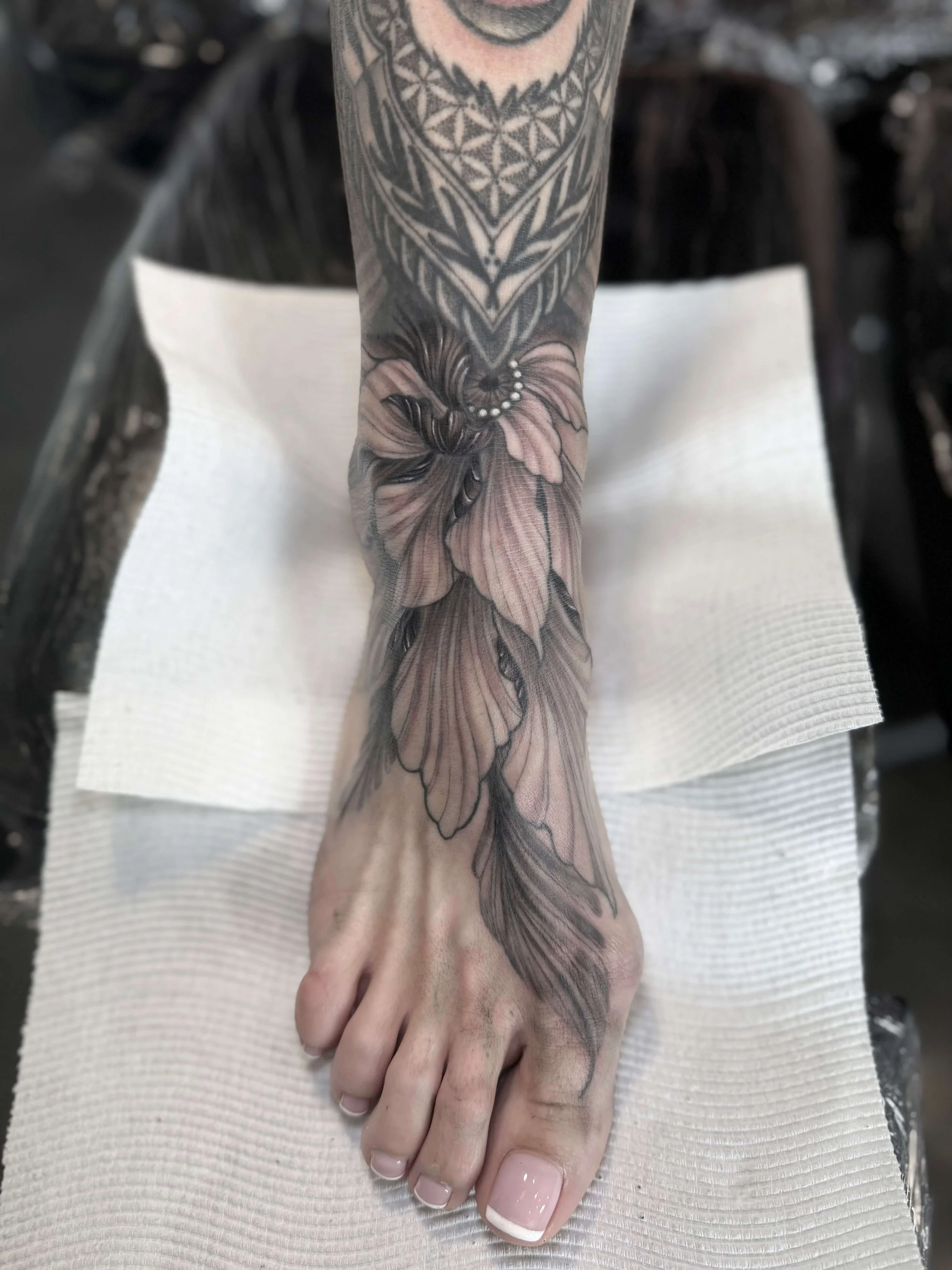Tattoo of a large flower with detailed petals and leaves on a person's ankle and lower leg, with geometric patterns above.