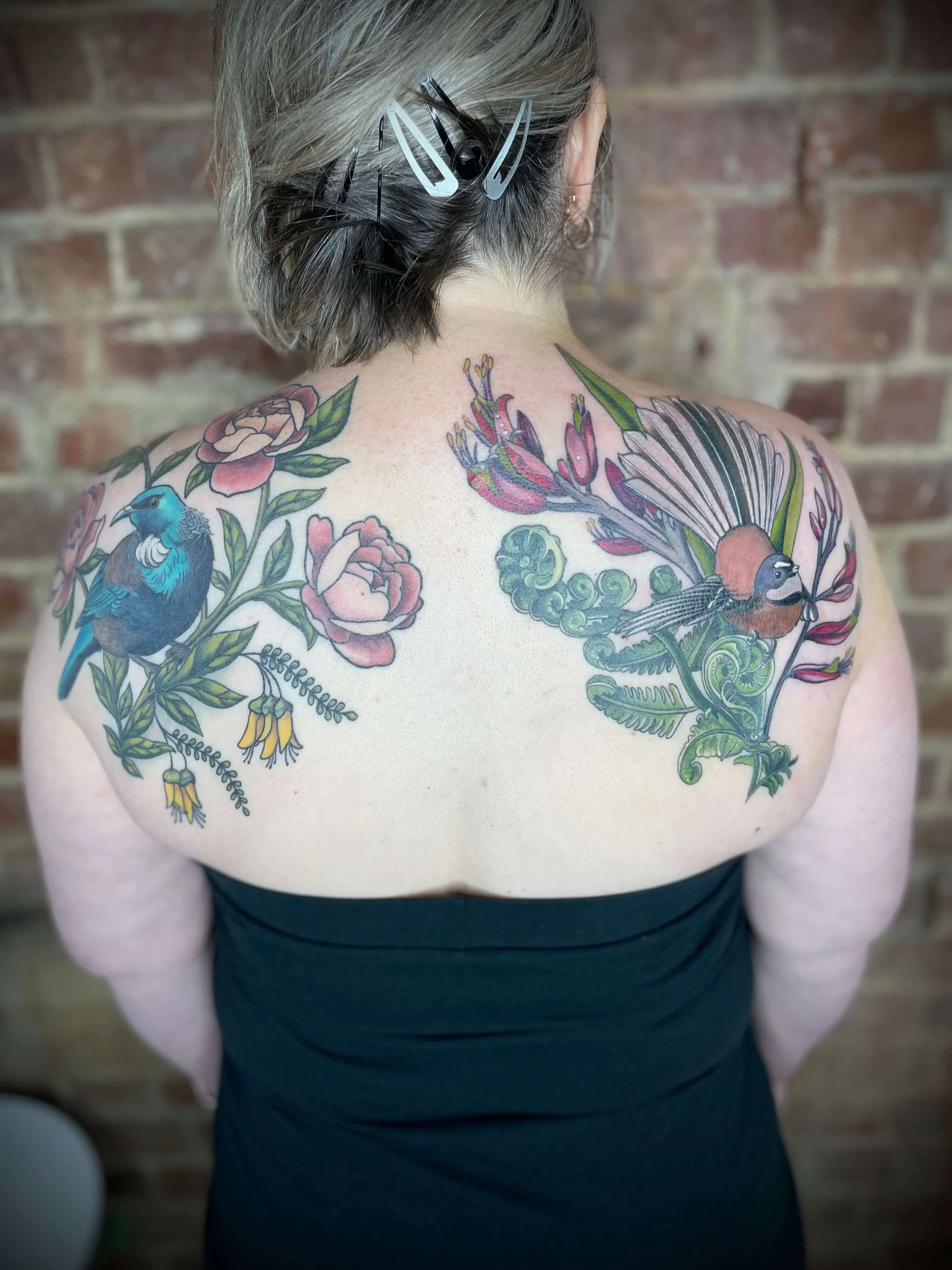 Colorful floral and bird tattoo on a person's back, with a brick wall in the background.