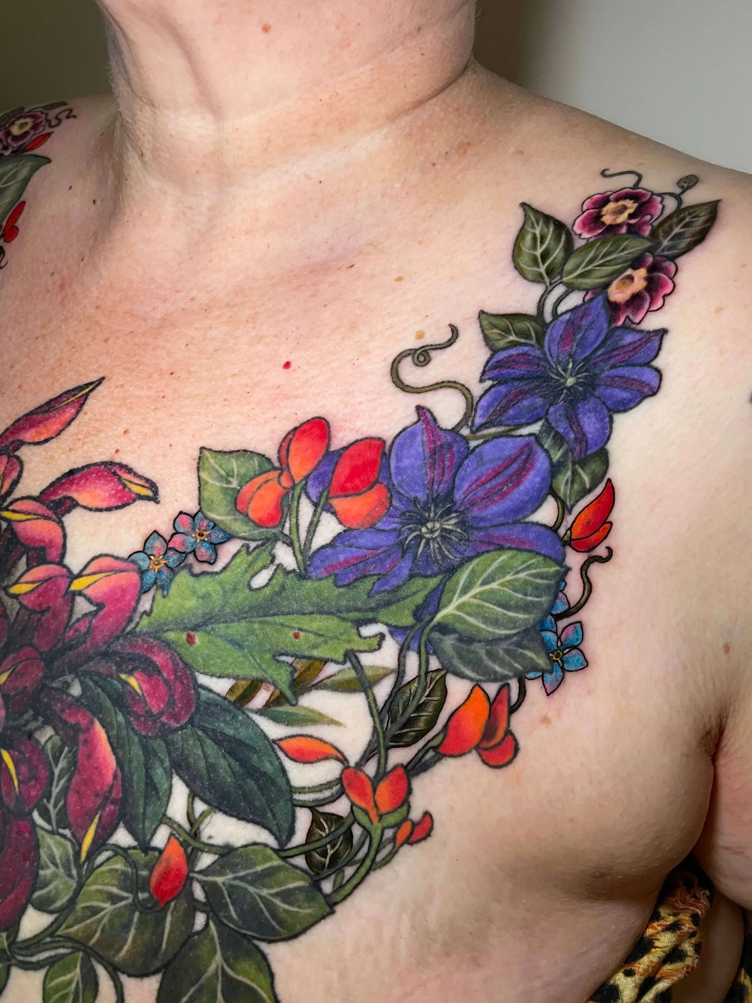 Colorful floral tattoo on person's chest and shoulder featuring purple, pink, red, and blue flowers with green leaves.