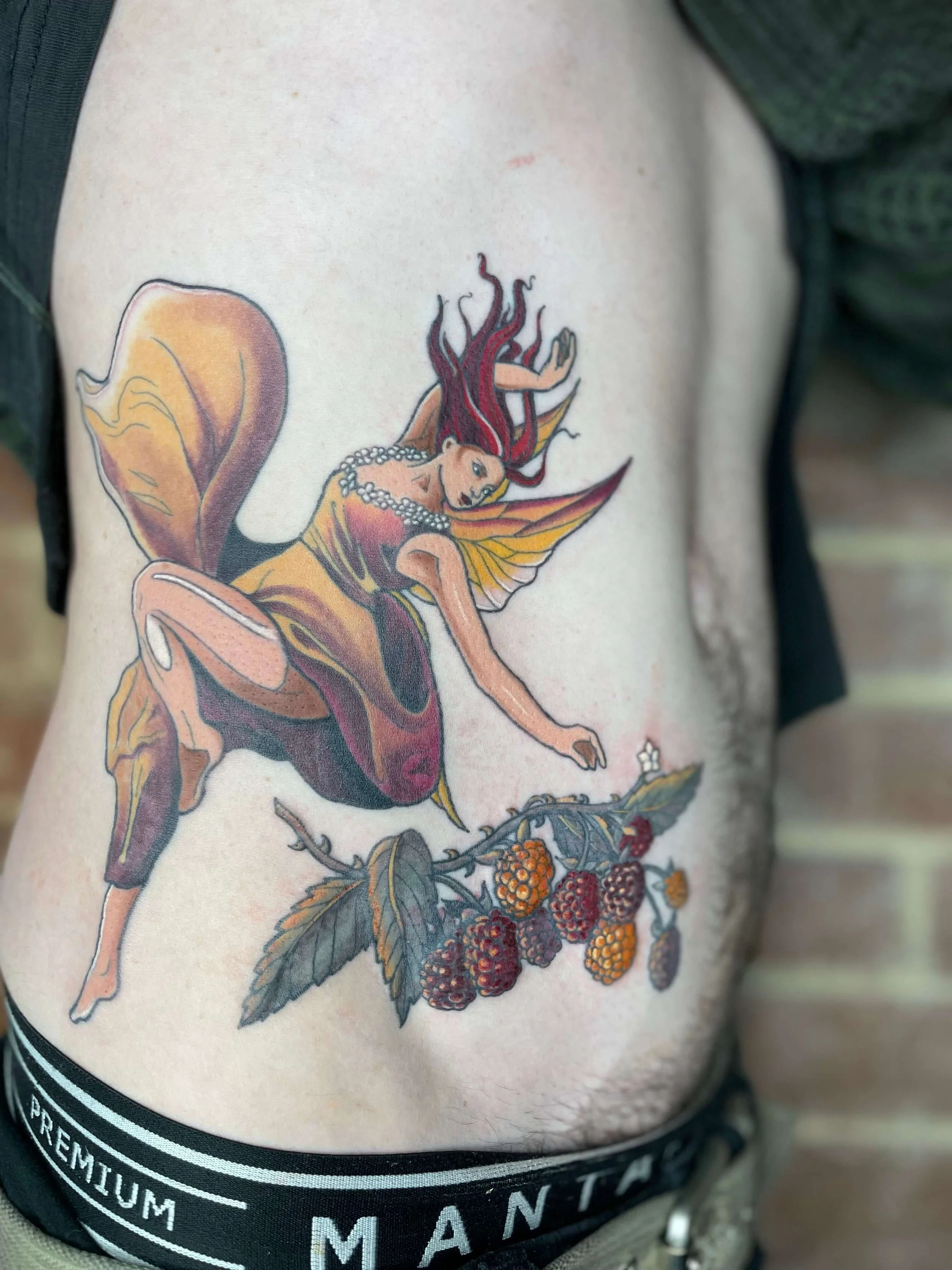 A tattoo of a fairy with red hair, butterfly wings, and a necklace, sitting on blackberry leaves.