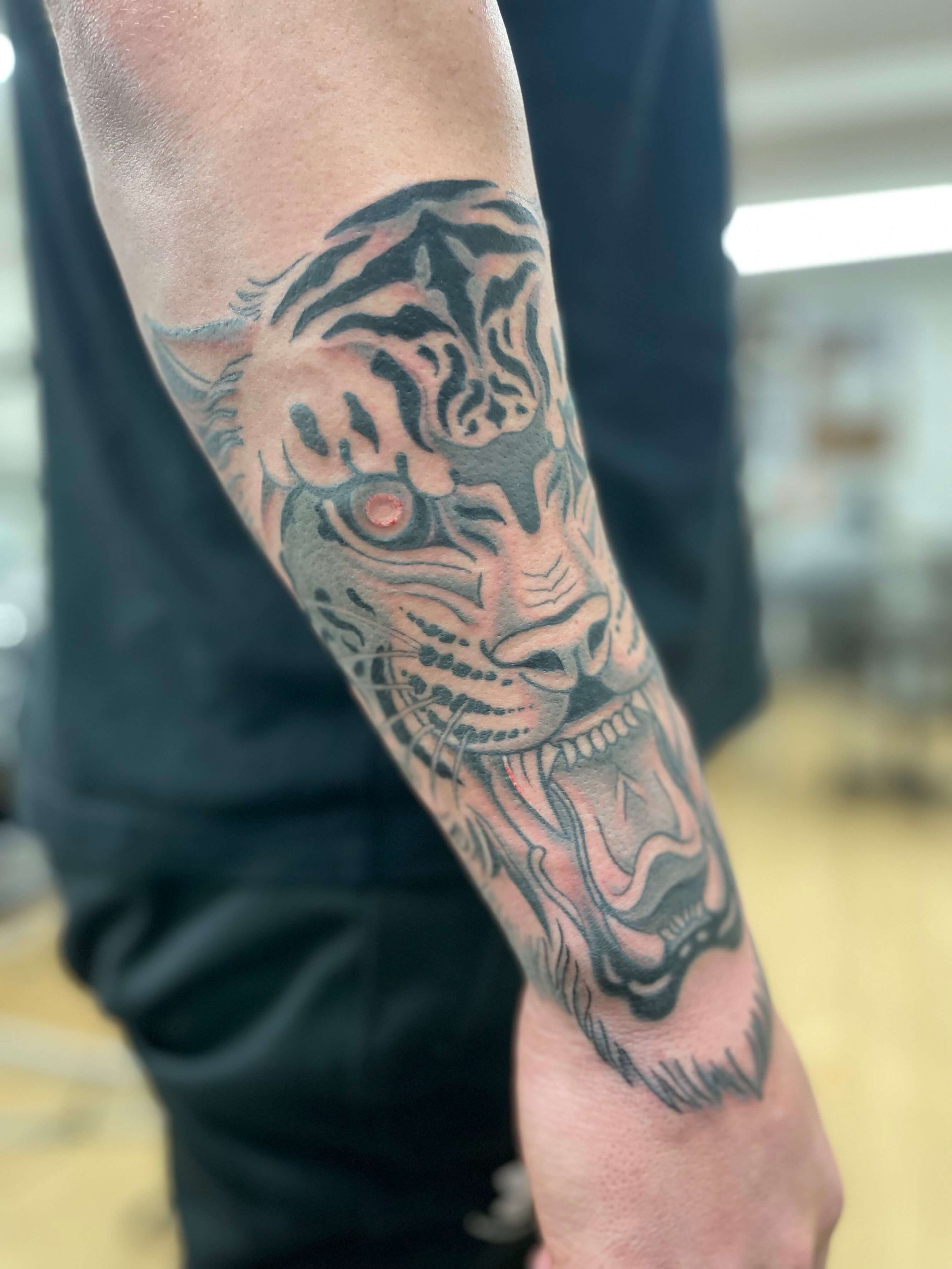 Tattoo of a tiger's face on a person's forearm, with the tiger's mouth open showing teeth and tongue.