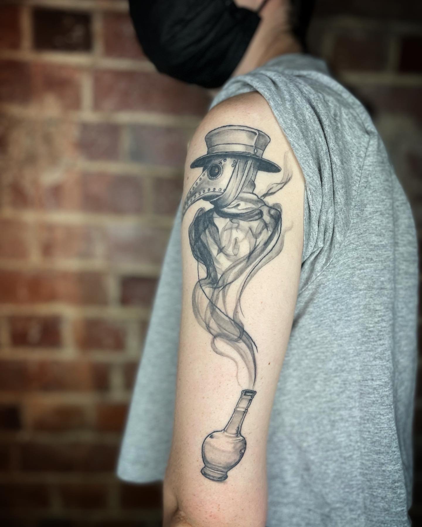 Tattoo of a plague doctor mask and hat, with smoke rising from a smoking pipe, on a person's upper arm.