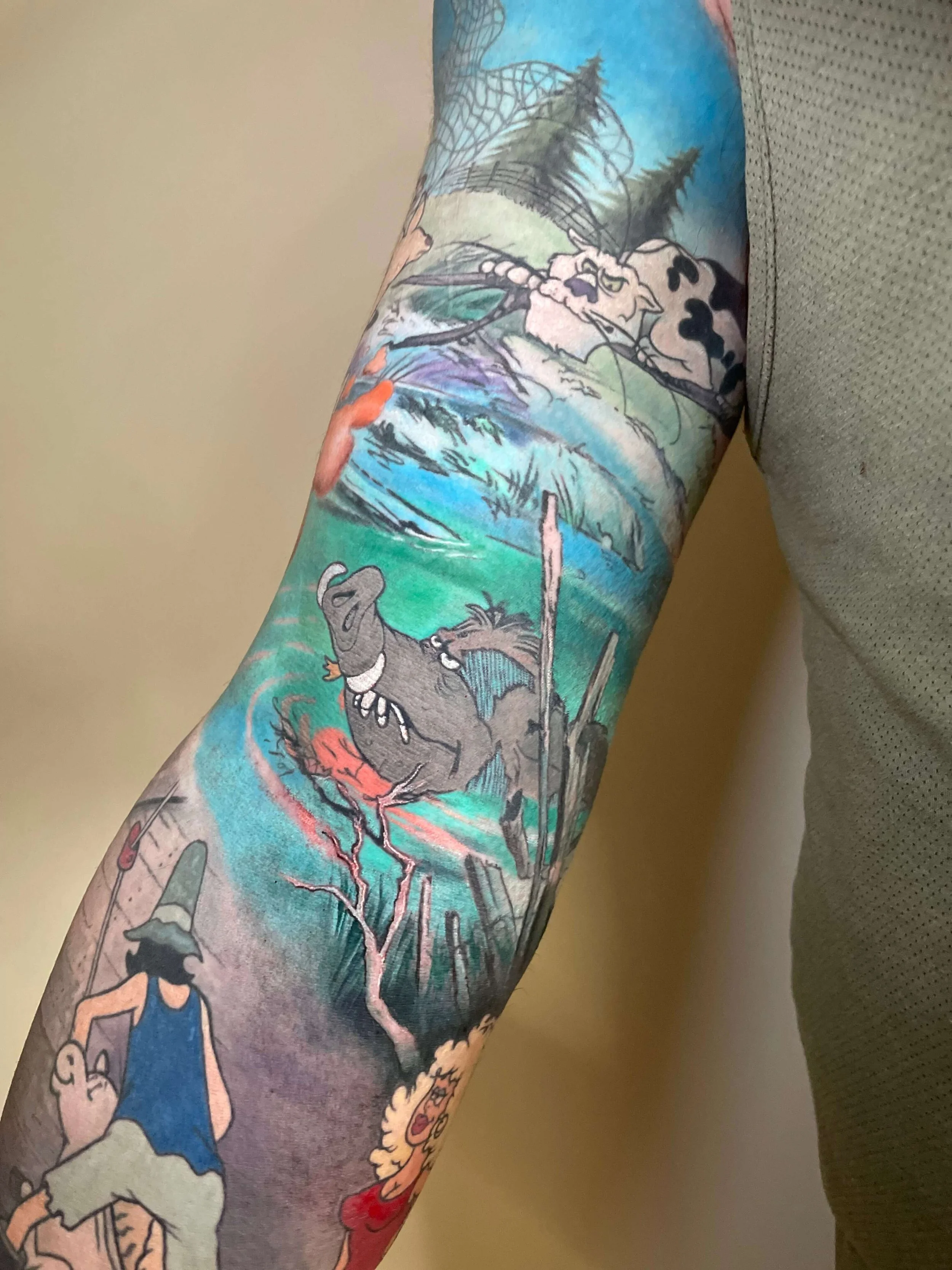 Colorful tattoo sleeve with various cartoon characters and scenes, including a bear, a person with a hat, and a landscape with trees and water.