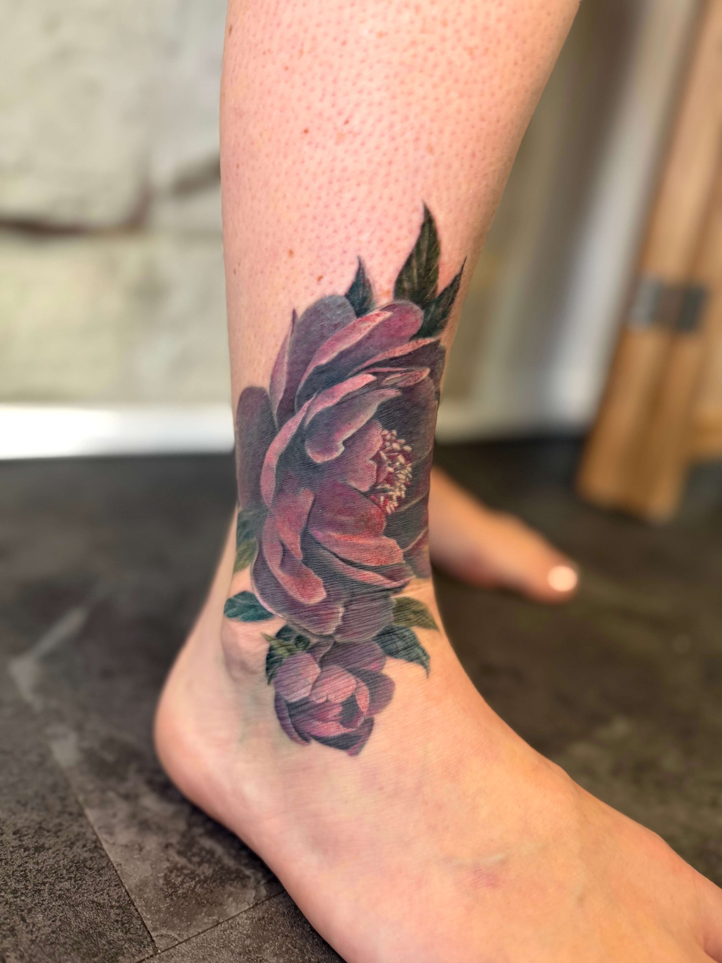 A close-up of a tattoo of a pink rose with green leaves on a person's ankle and lower leg.