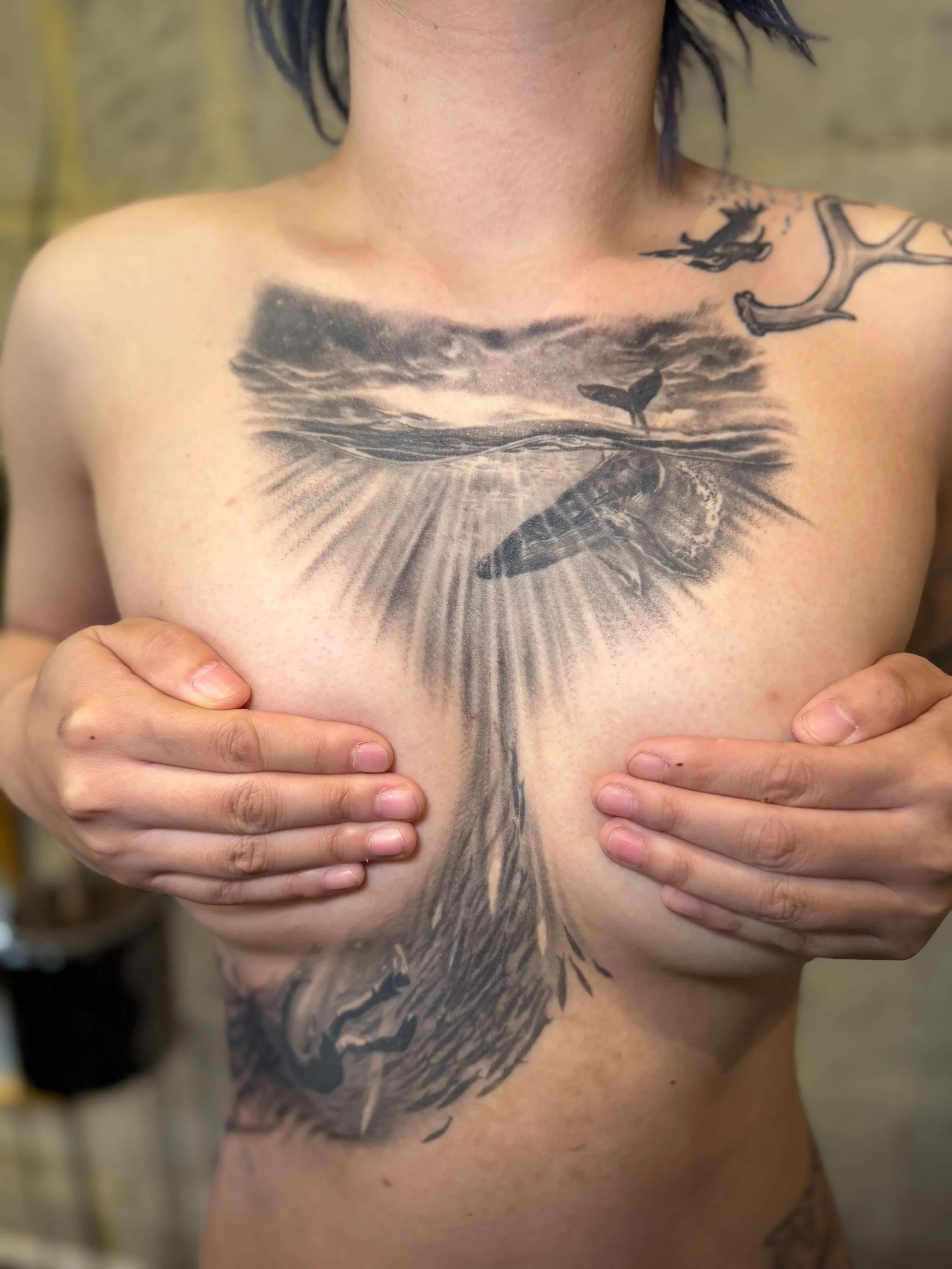 A person with black tattoos on their chest and shoulders, holding their breasts with their hands. The main tattoo features a detailed black and gray whale appearing in a cloudy sky with sun rays and ocean waves below.
