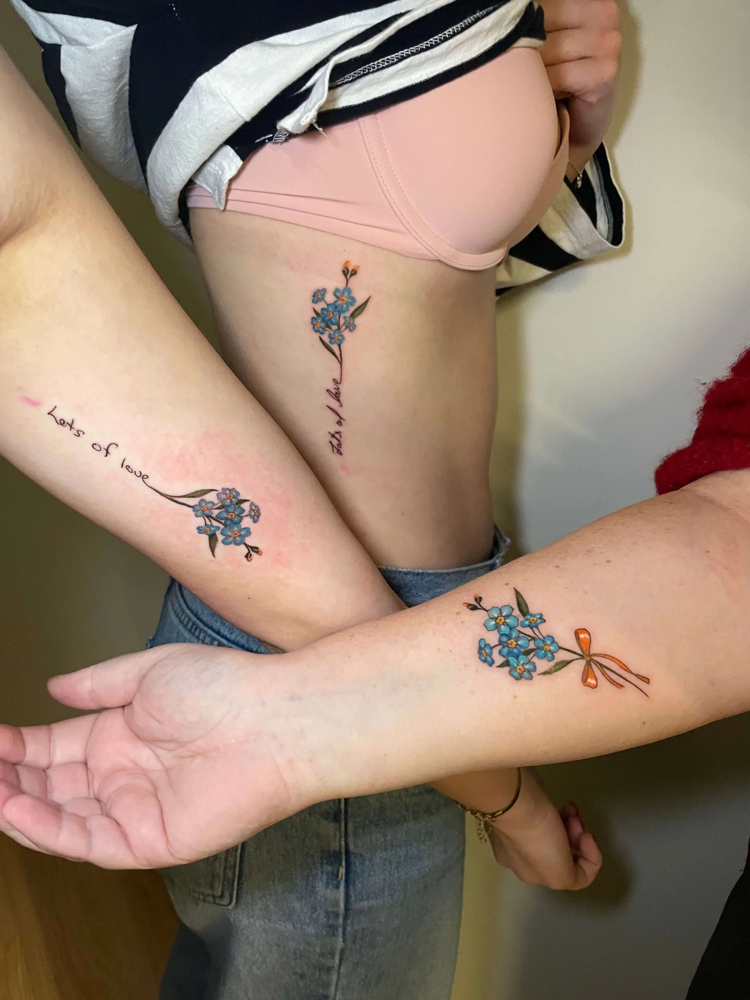 Three people with matching tattoos of blue flowers and orange bows on their arms and side, with the words 'Let’s of love' and 'tsh Mashi' written beside the tattoos.