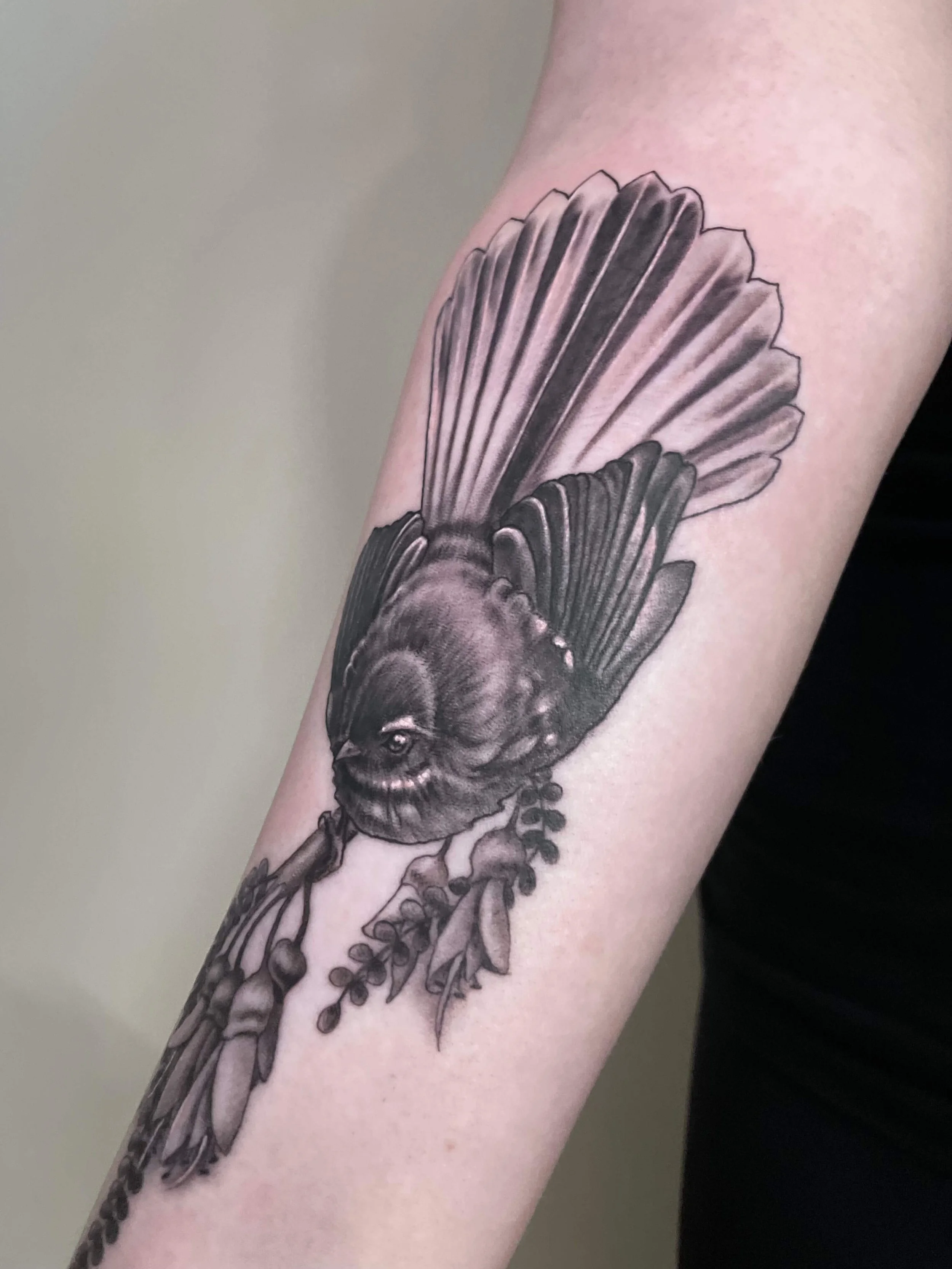 Black and gray realistic tattoo of a bird perched on a branch with leaves on a person's arm.