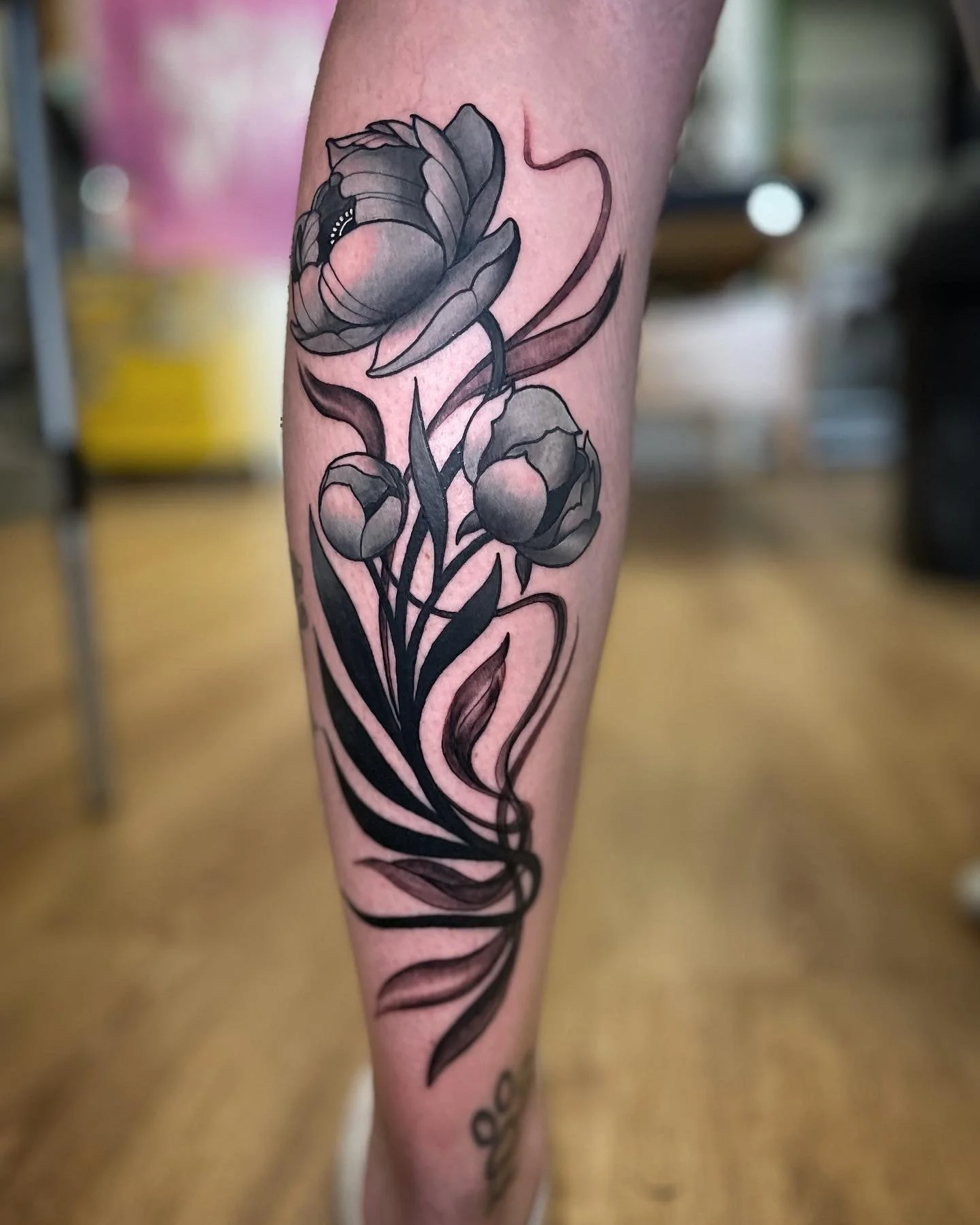 Black and gray floral tattoo featuring a large open flower, two smaller buds, and flowing leaves on a person's calf.