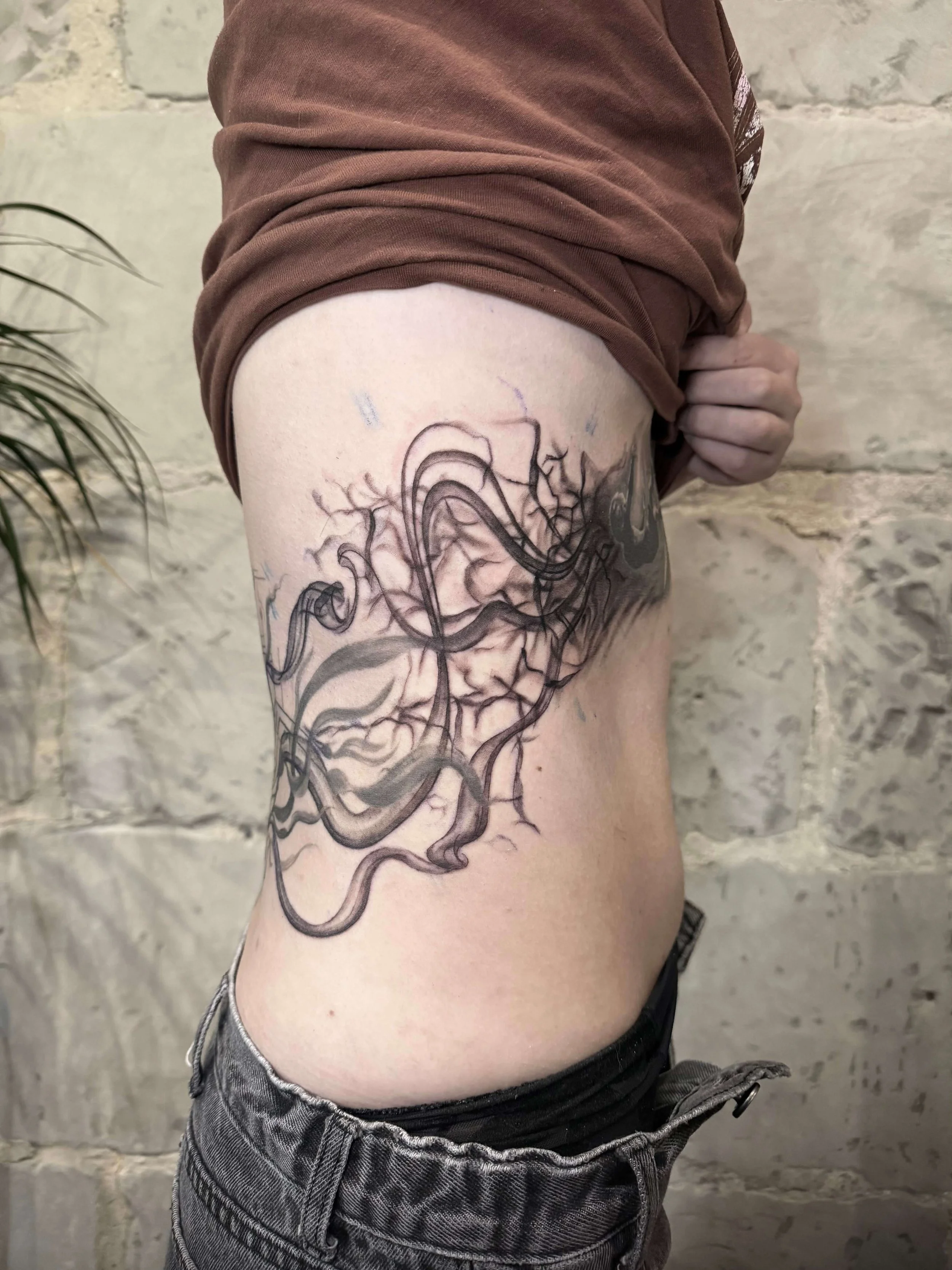 A person lifting their shirt to reveal a detailed black and gray tattoo of a woman's face intertwined with abstract, flowing lines and crackle-like patterns on their side.