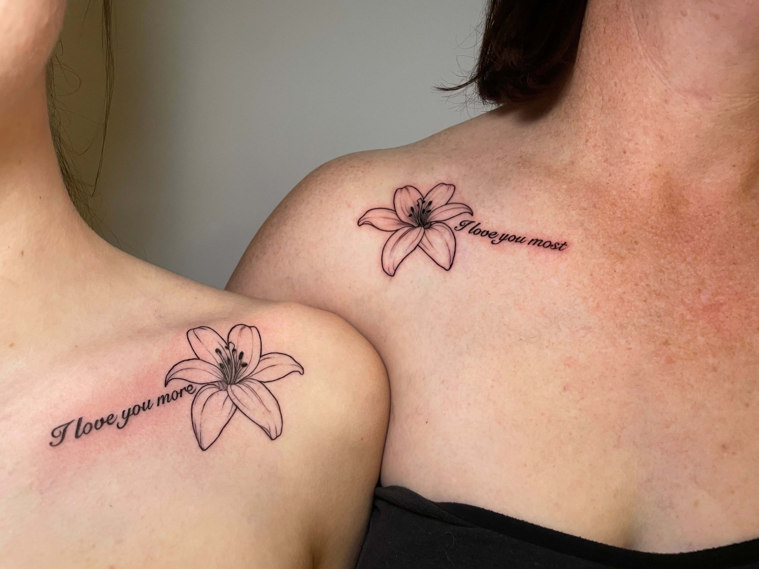 Close-up of two women's shoulders with matching tattoos of lily flowers and the phrase 'I love you most' in cursive.